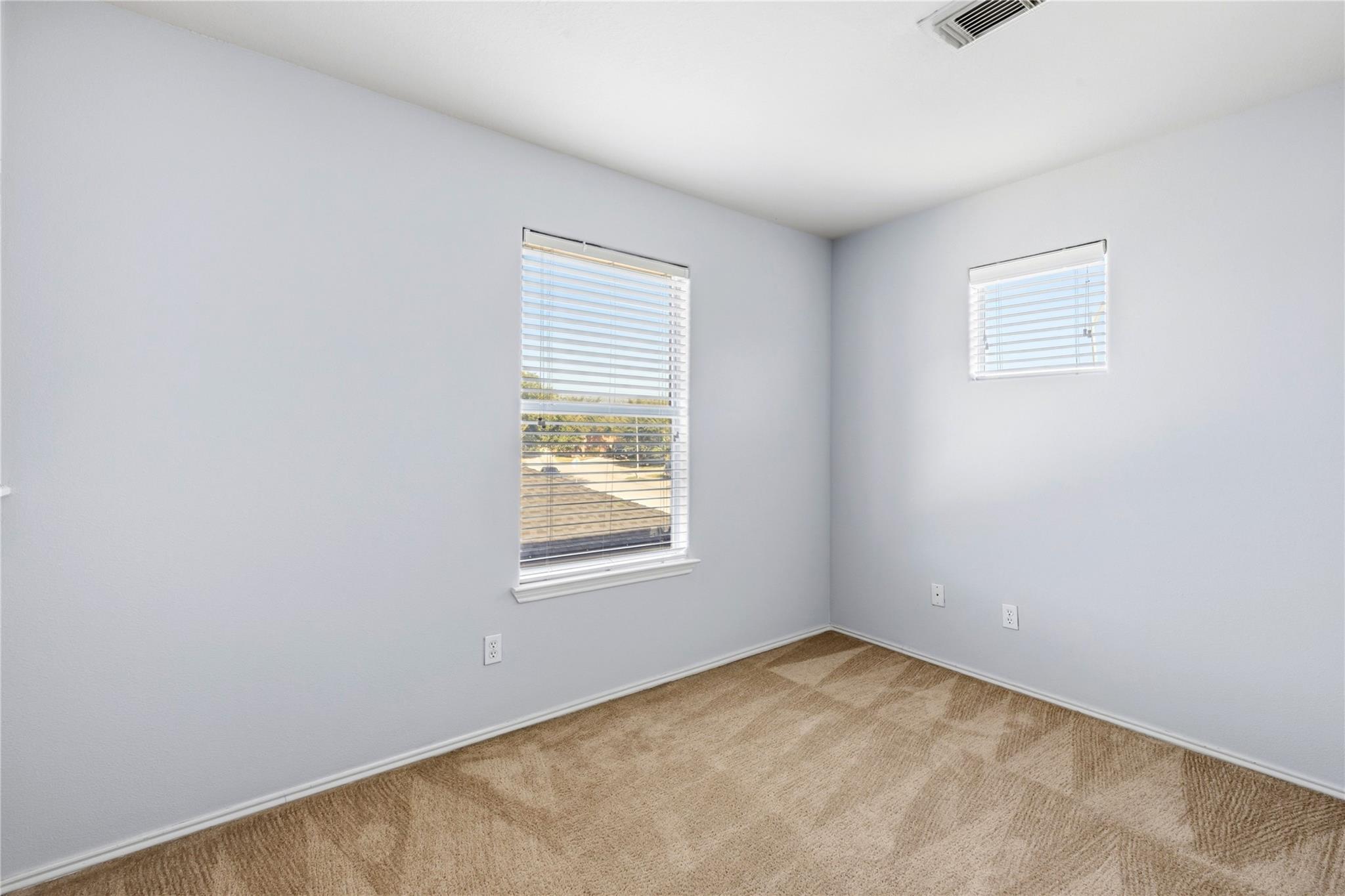 Empty carpeted room with pale gray walls and two windows with white blinds.