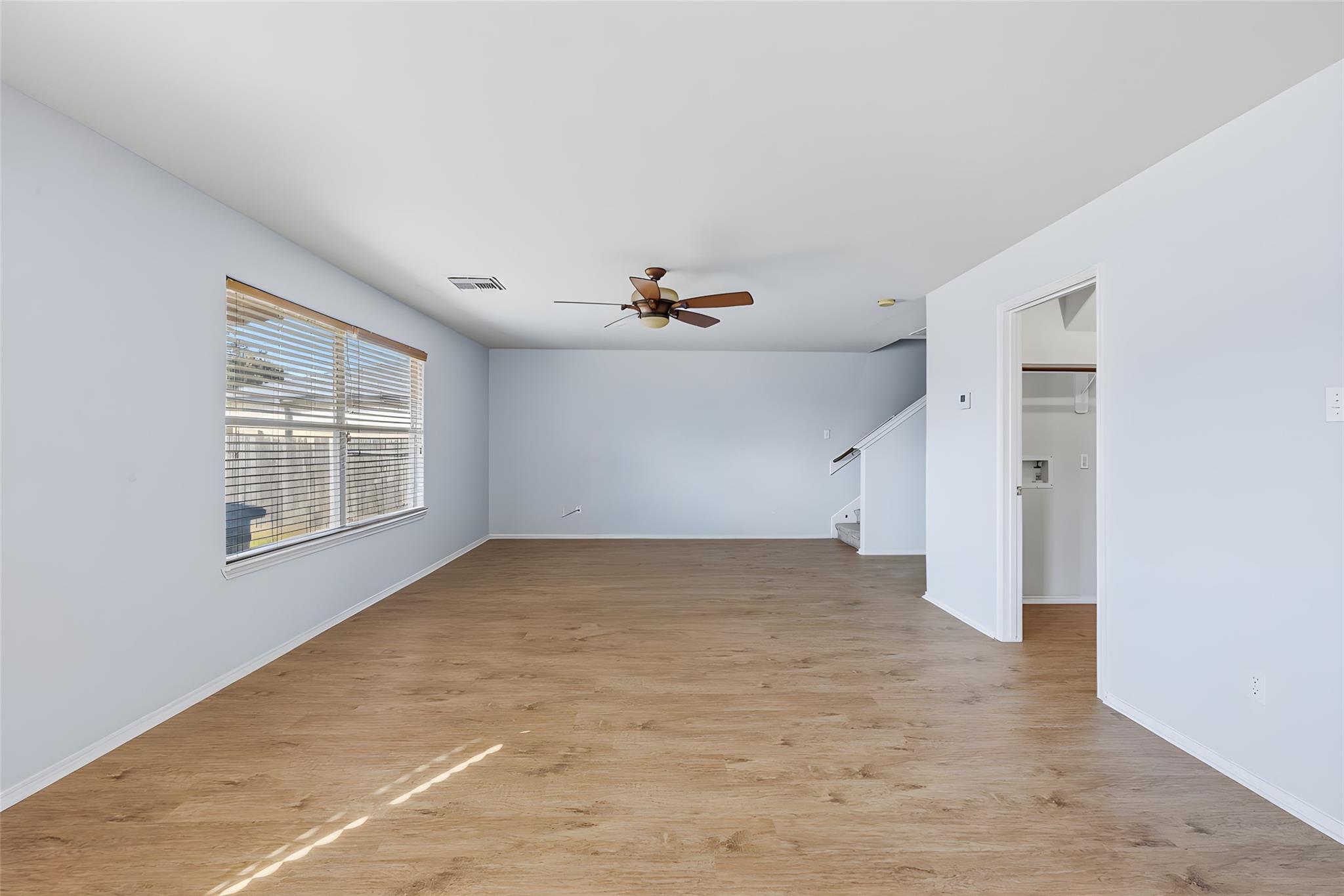 Empty living room with wood-look floors, a ceiling fan, and a large window with blinds.