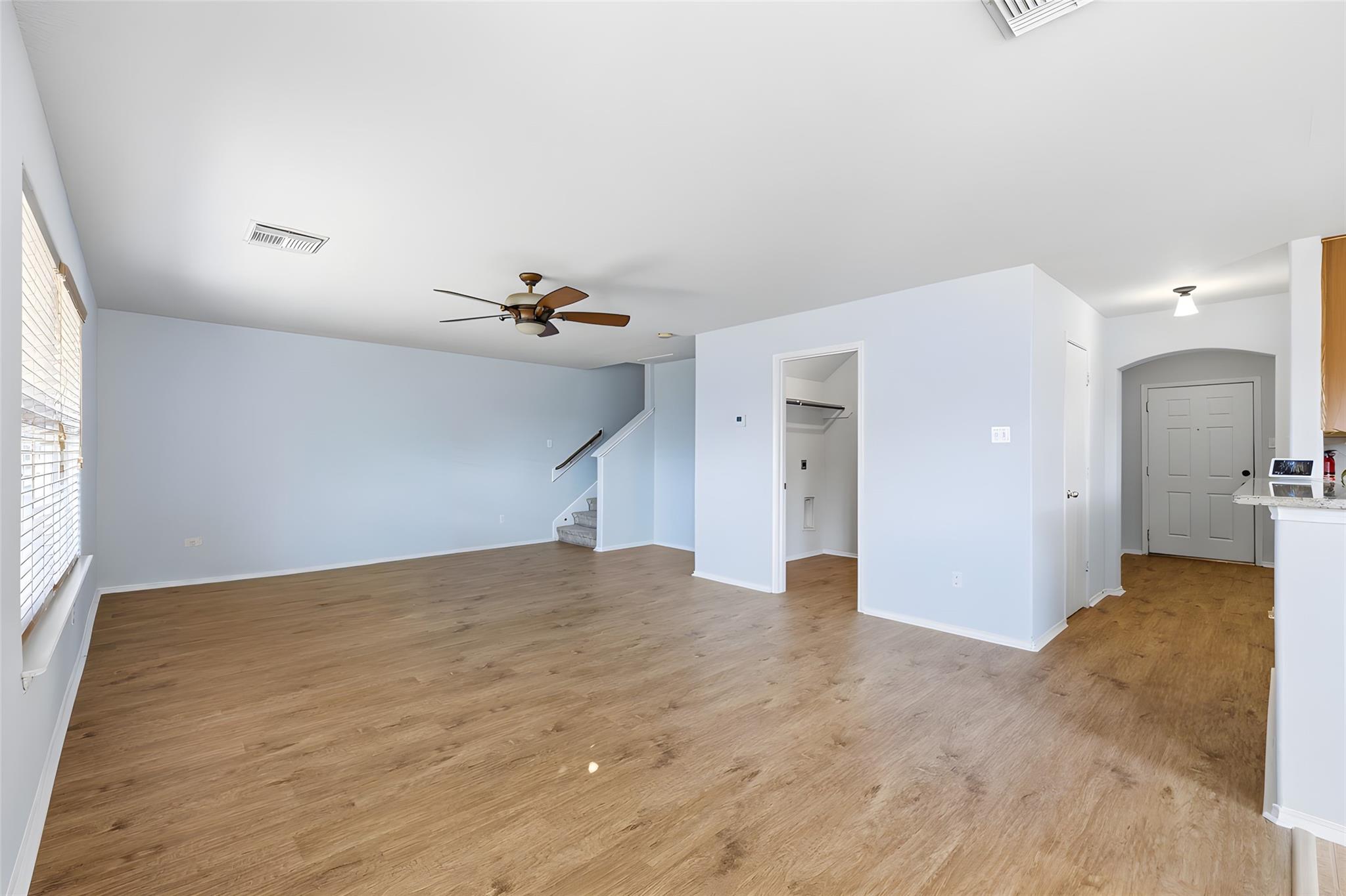 Empty living room with light wood floors, a ceiling fan, staircase, and front entry.