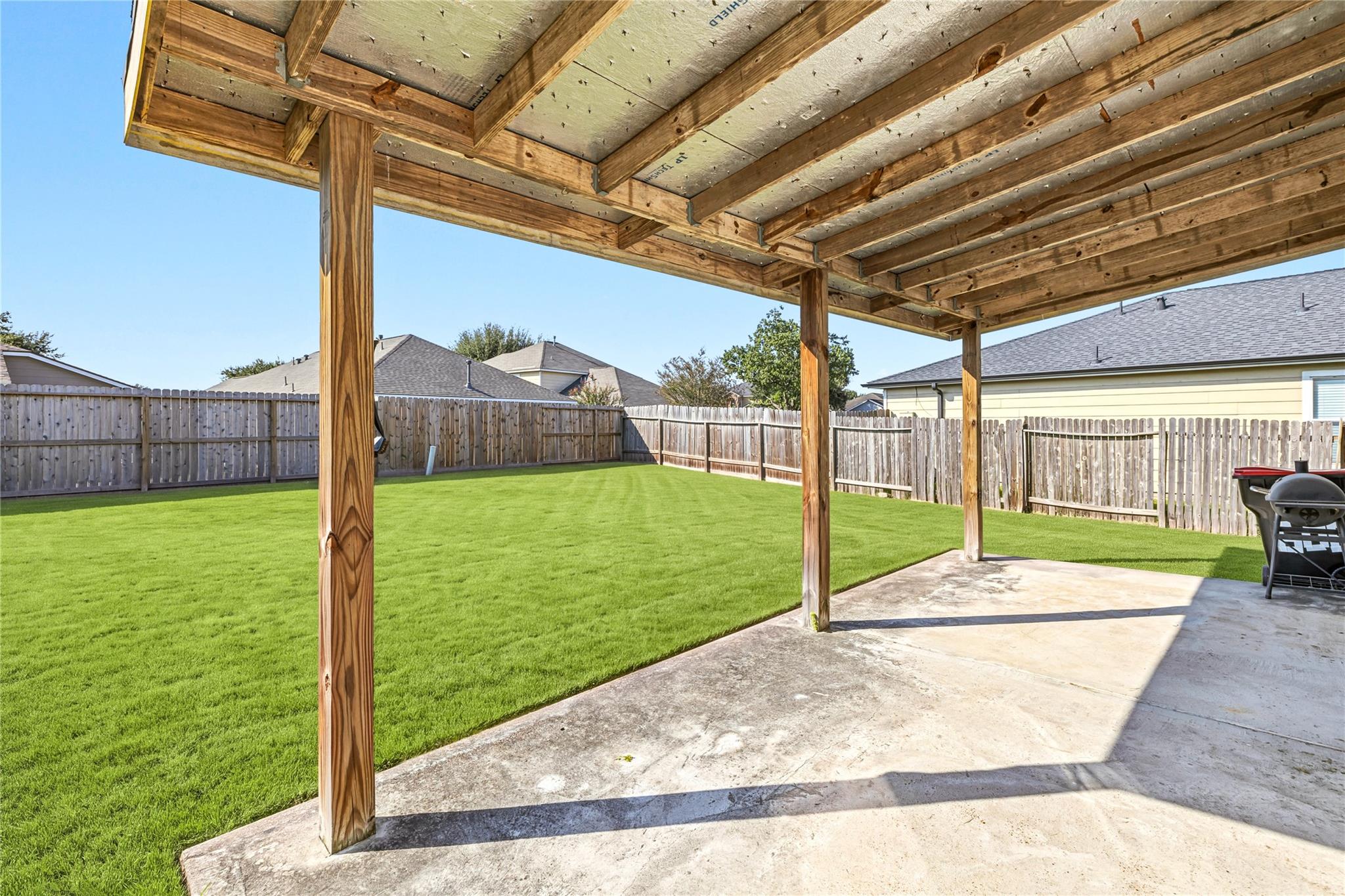 Covered concrete patio with exposed timber framing beside a fenced, neatly mowed backyard lawn.