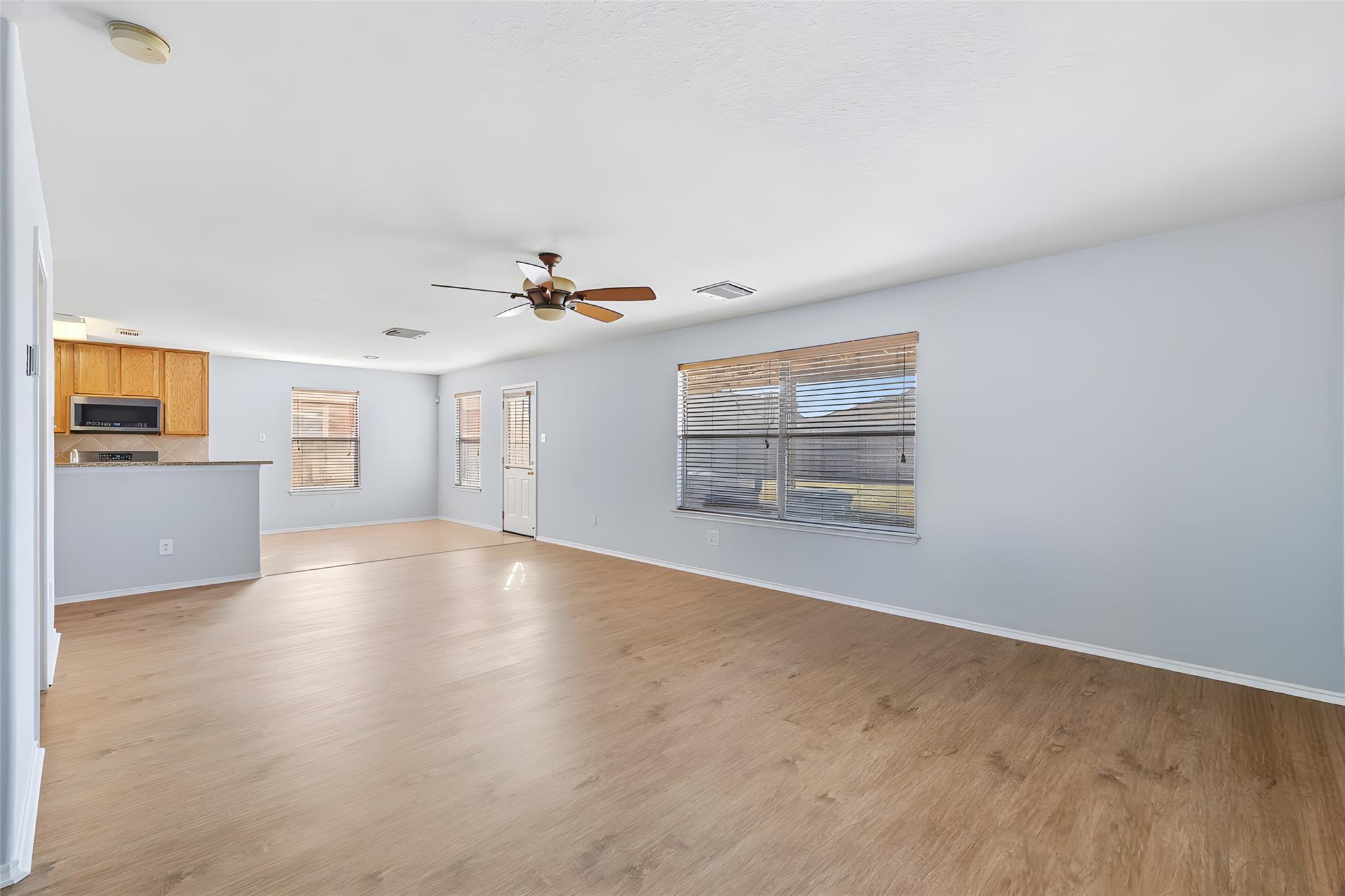 Empty living room with light wood floors, a ceiling fan, wide blinded windows, and an open kitchen.