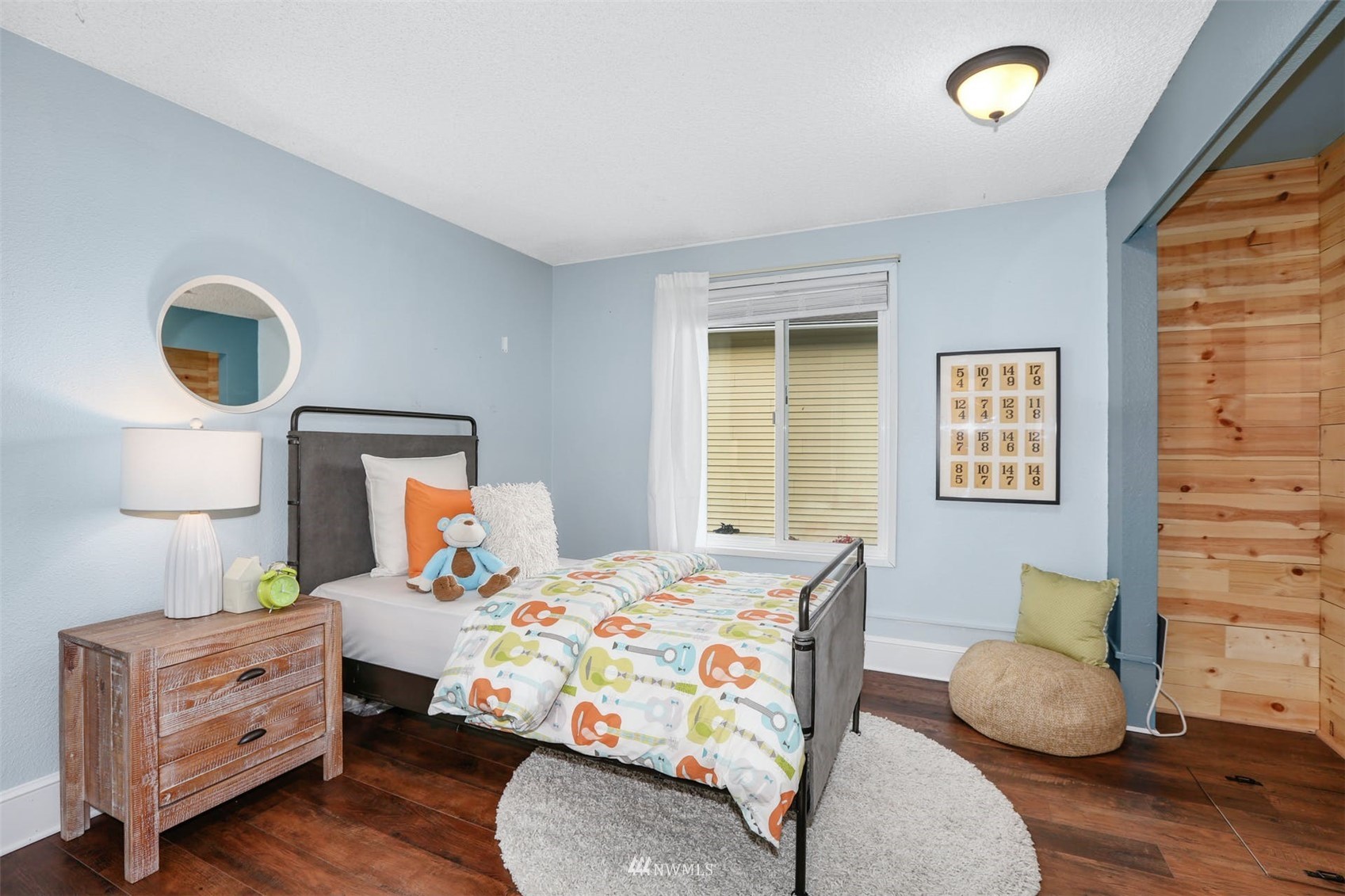 Furnished bedroom with colorful guitar-print bedding, pale blue walls, and dark wood floors.