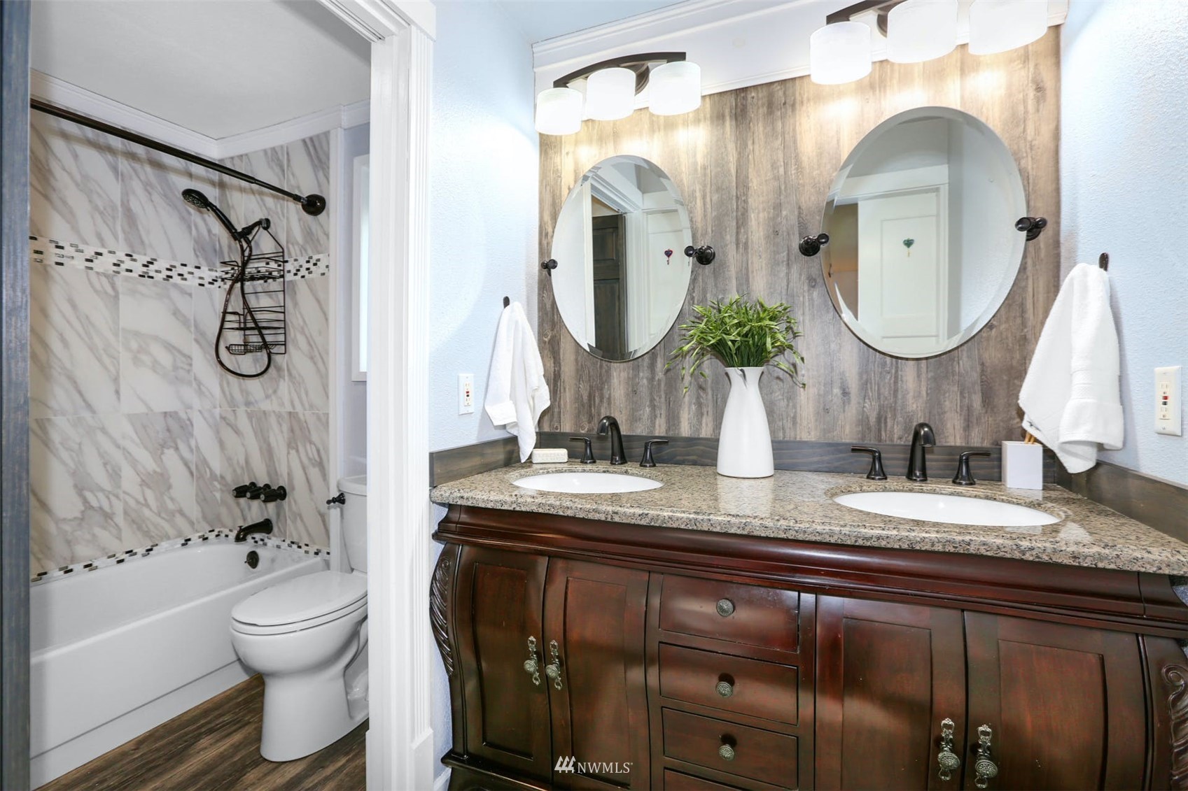 Bathroom with a dark double vanity, two oval mirrors, and a marble-look tiled tub and shower.