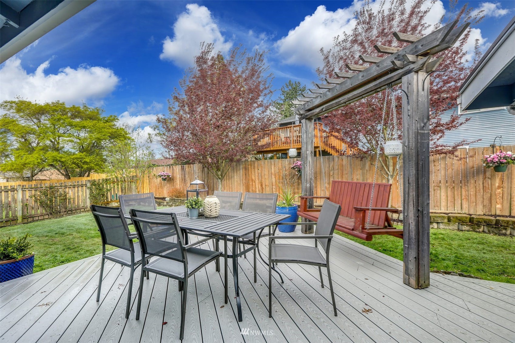 Backyard deck with a patio dining set, wooden pergola swing, fenced lawn, and flowering trees.
