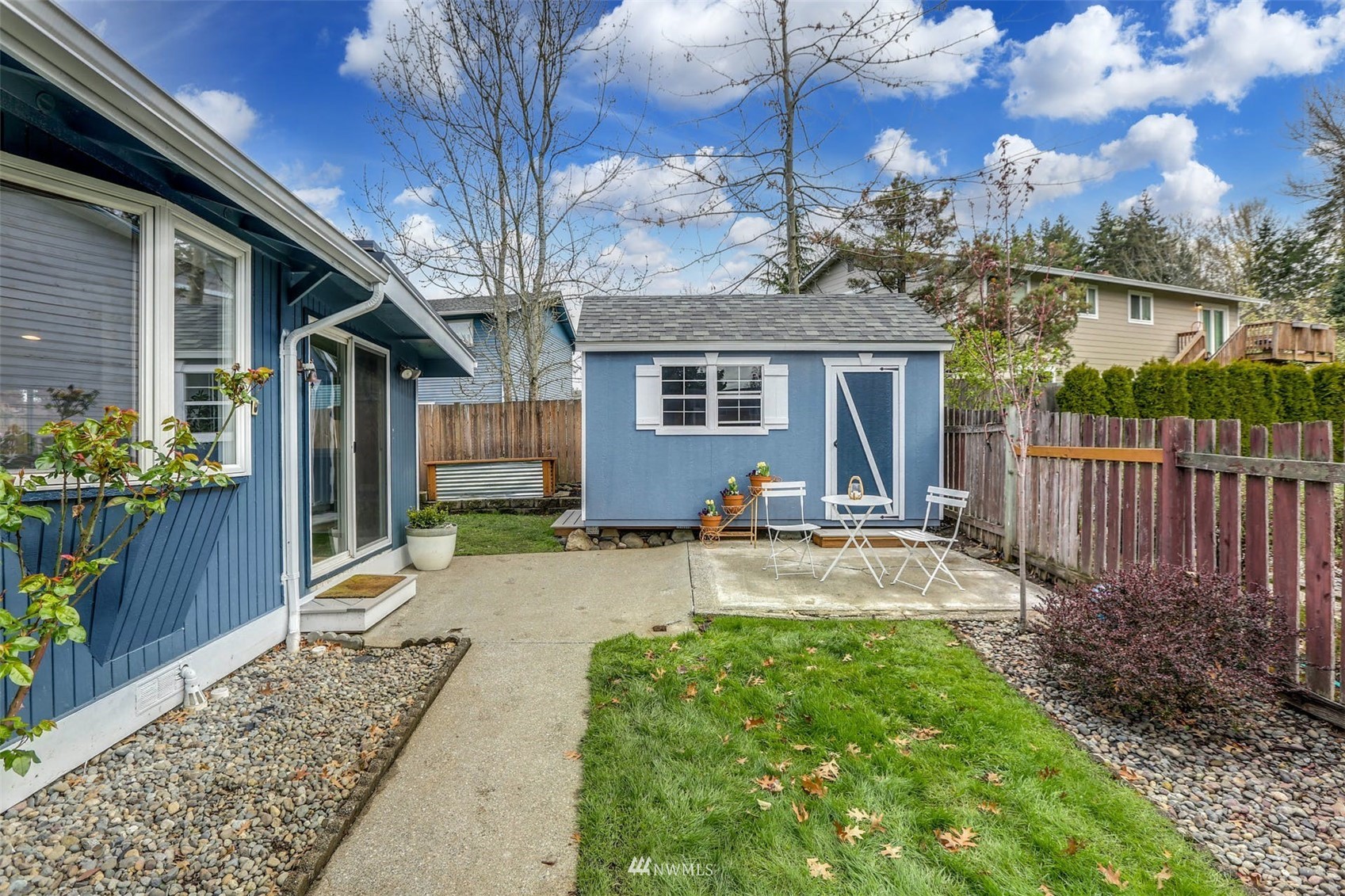 Backyard with a blue shed, fenced lawn, and concrete patio with outdoor chairs.