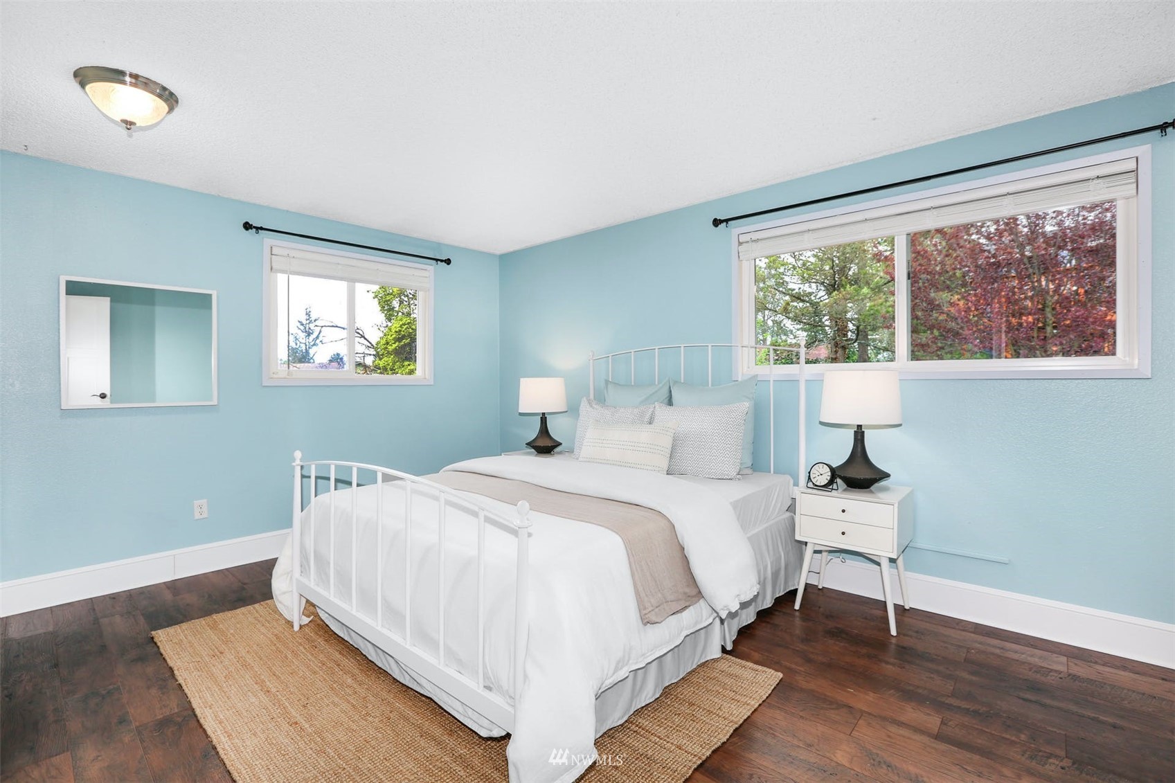 Staged bedroom with pale blue walls, a white metal bed, and two windows overlooking trees.