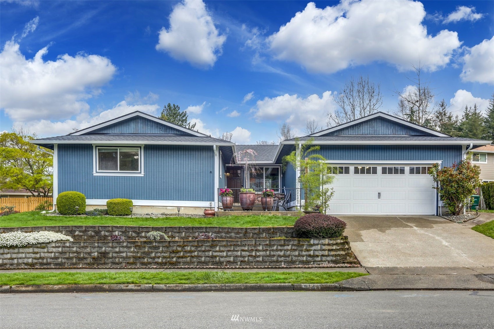 Blue single-story home with a white double garage door, potted flowers, and a tiered stone retaining wall.