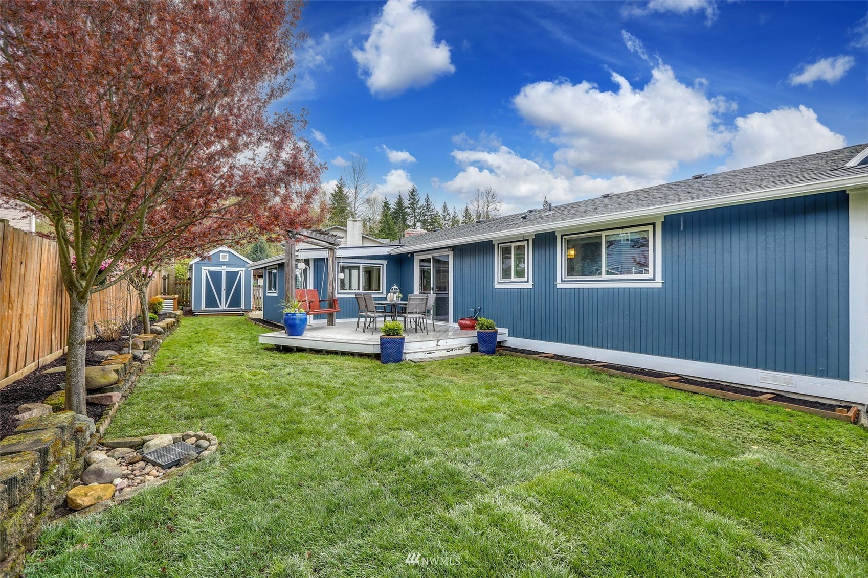 Blue home and grassy backyard with a furnished deck, blue shed, and autumn tree.
