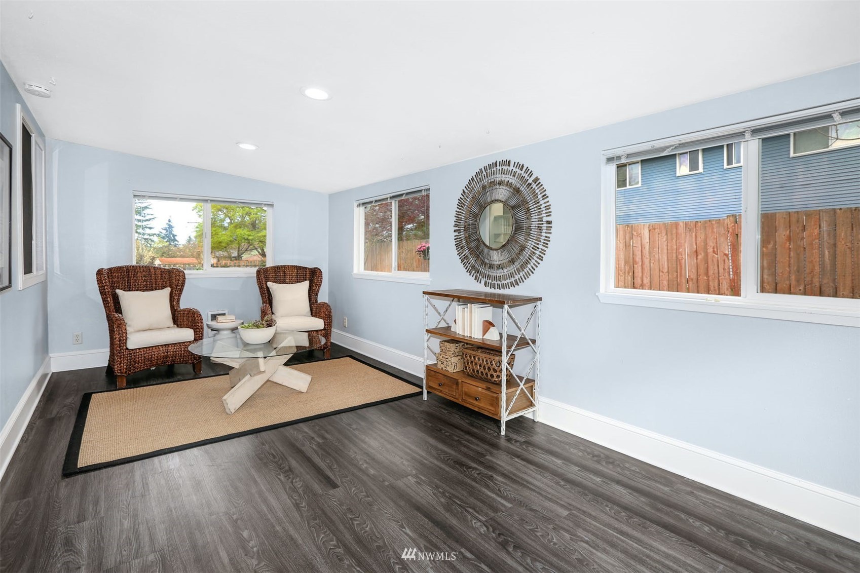 Staged living room with wicker armchairs, a woven rug, and windows facing a fenced yard.