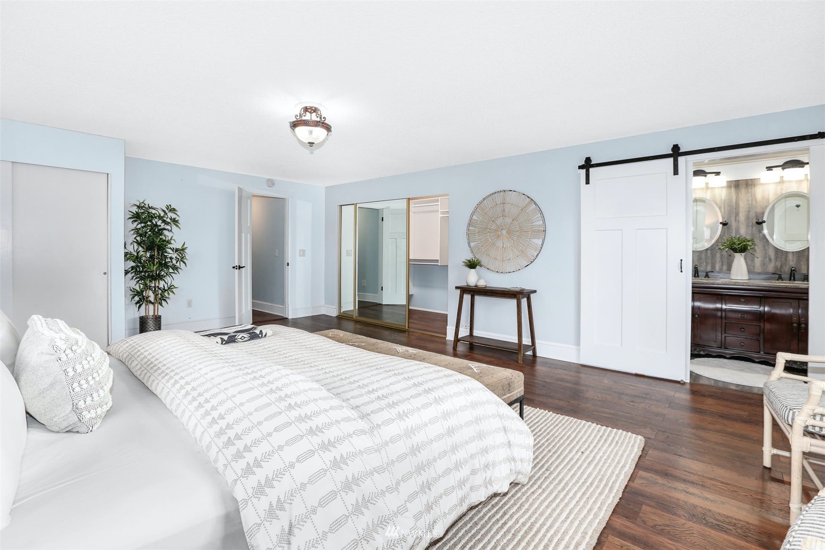 Staged bedroom with a patterned white bed, mirrored closet doors, and a barn door opening to the bathroom.