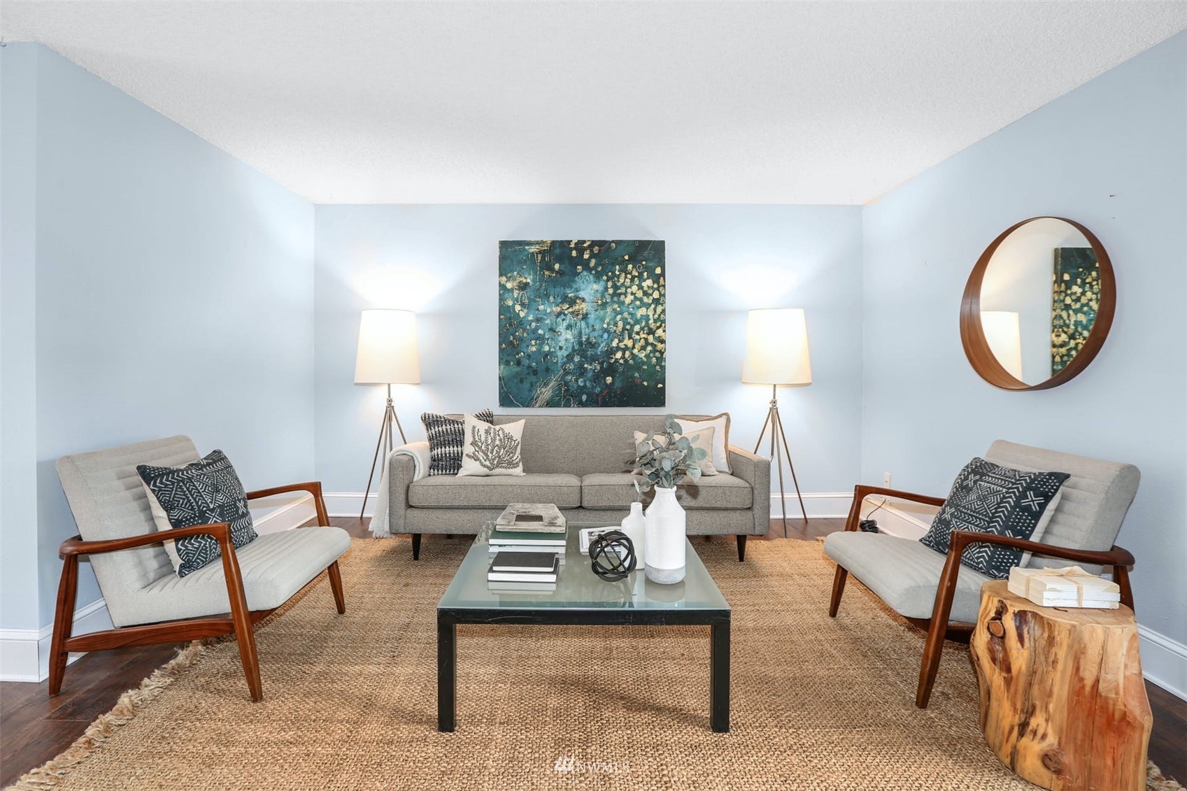 Staged living room with pale blue walls, gray seating, a glass coffee table, and a large woven rug.