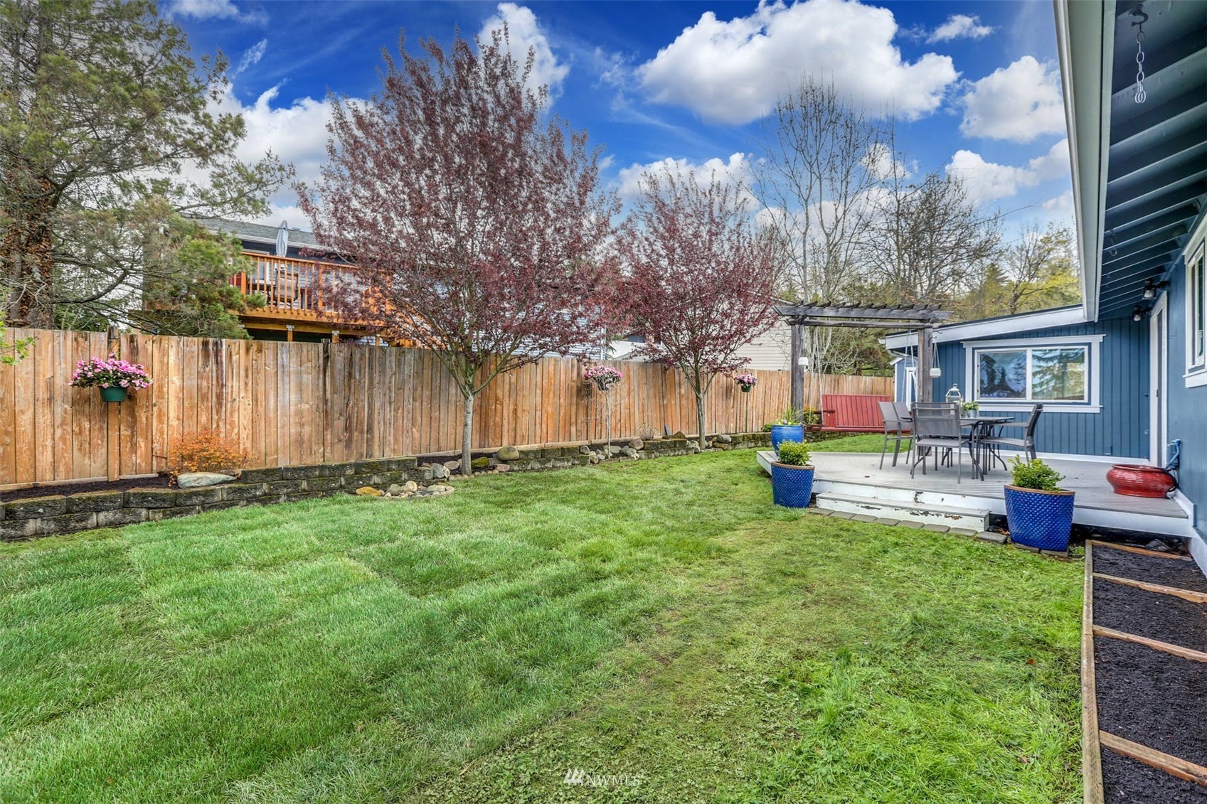 Fenced backyard with a broad lawn, flowering trees, and a raised patio with outdoor dining furniture.