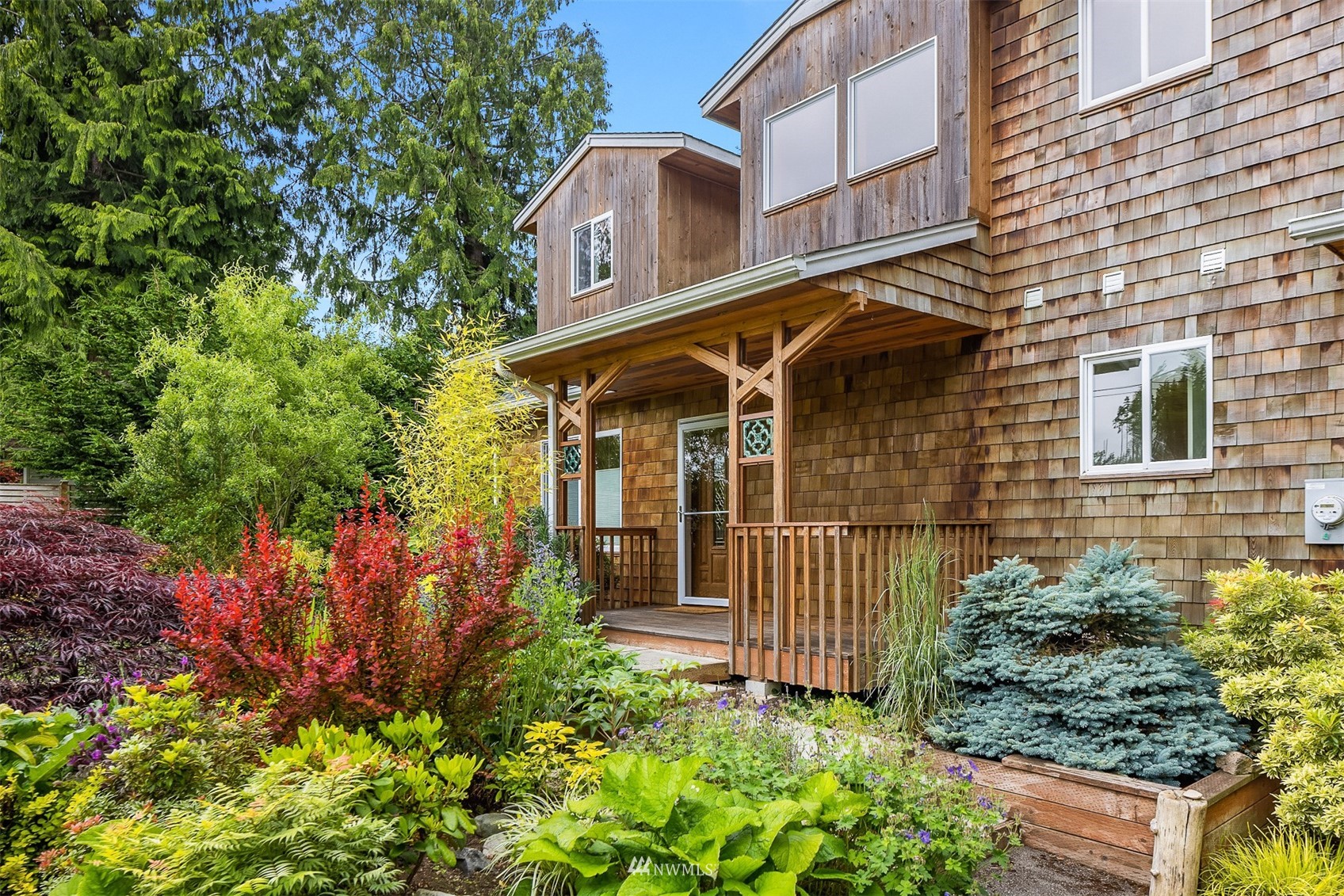 Cedar-shingle house with a covered porch, framed by colorful garden plants and evergreen trees.