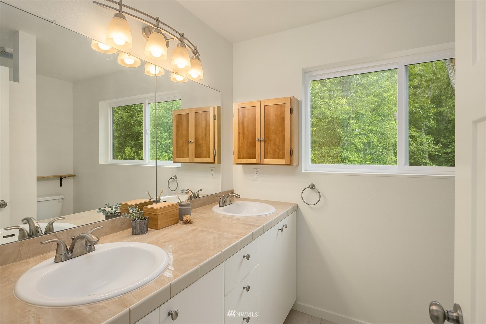 Bathroom with a tiled double-sink vanity, mirrored wall, wooden cabinets, and a wide window facing trees.