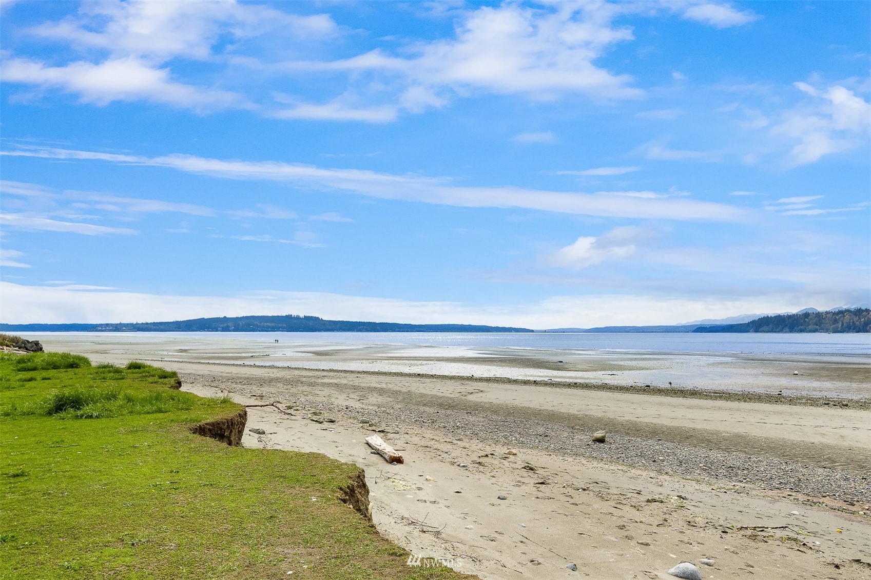 Low-tide beach with grassy shoreline, calm water, and distant wooded shores.