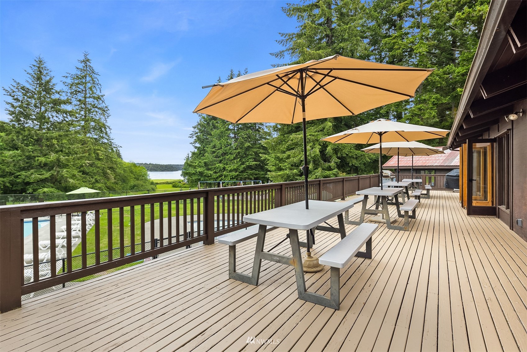 Wood deck with umbrella-shaded picnic tables overlooking a lawn, pool, trees, and water.