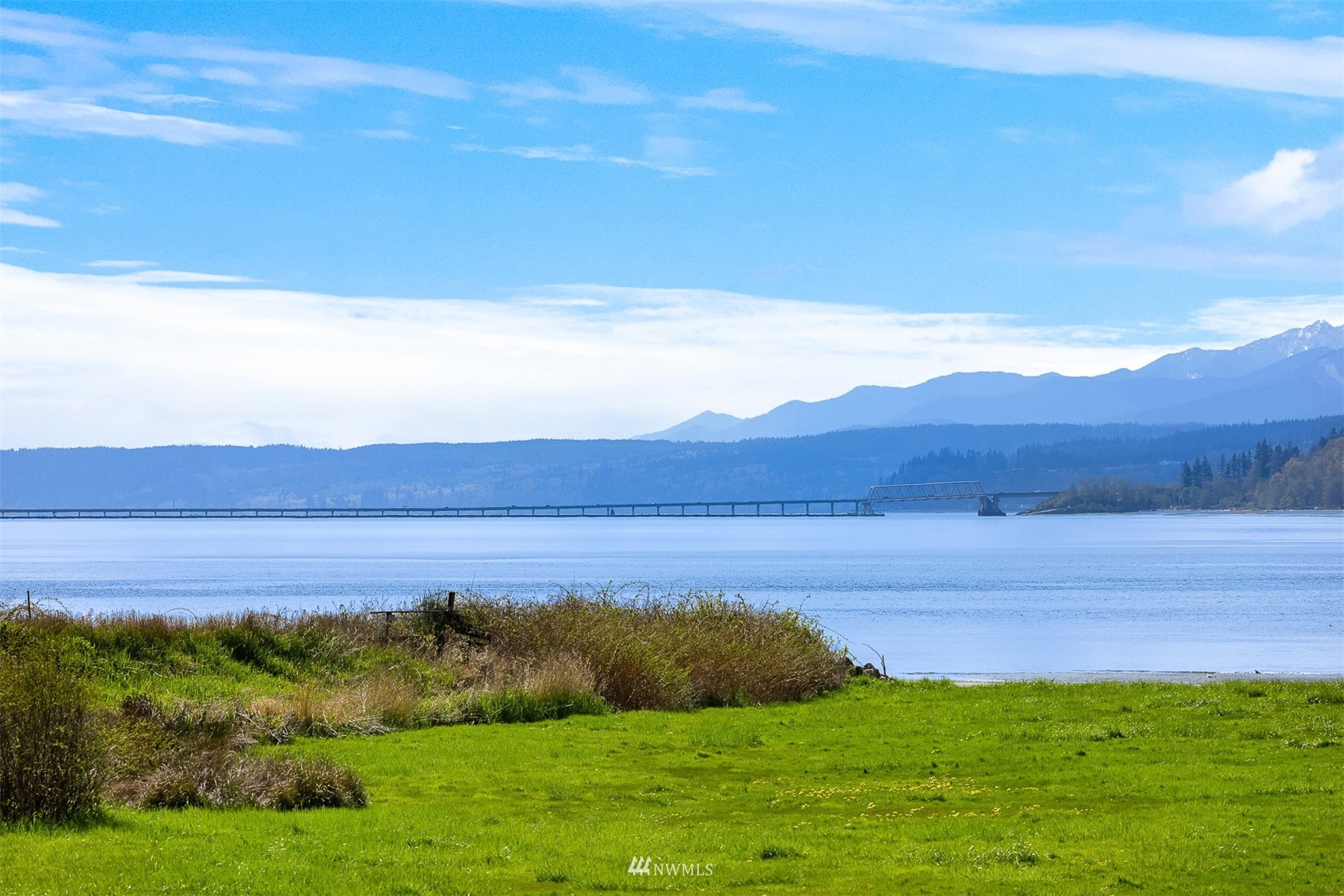 Waterfront view across a grassy shore to a long bridge and distant mountains.