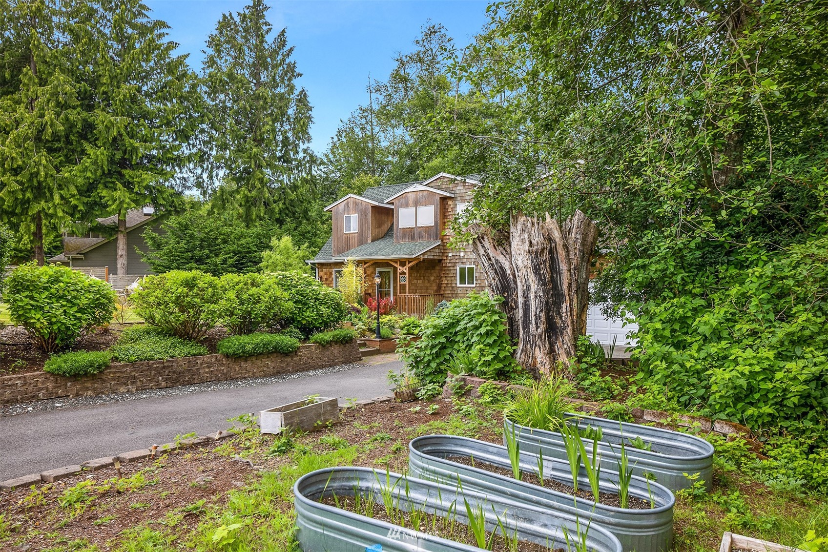 Cedar-shingle home amid mature trees, with raised metal garden beds and a large tree stump.