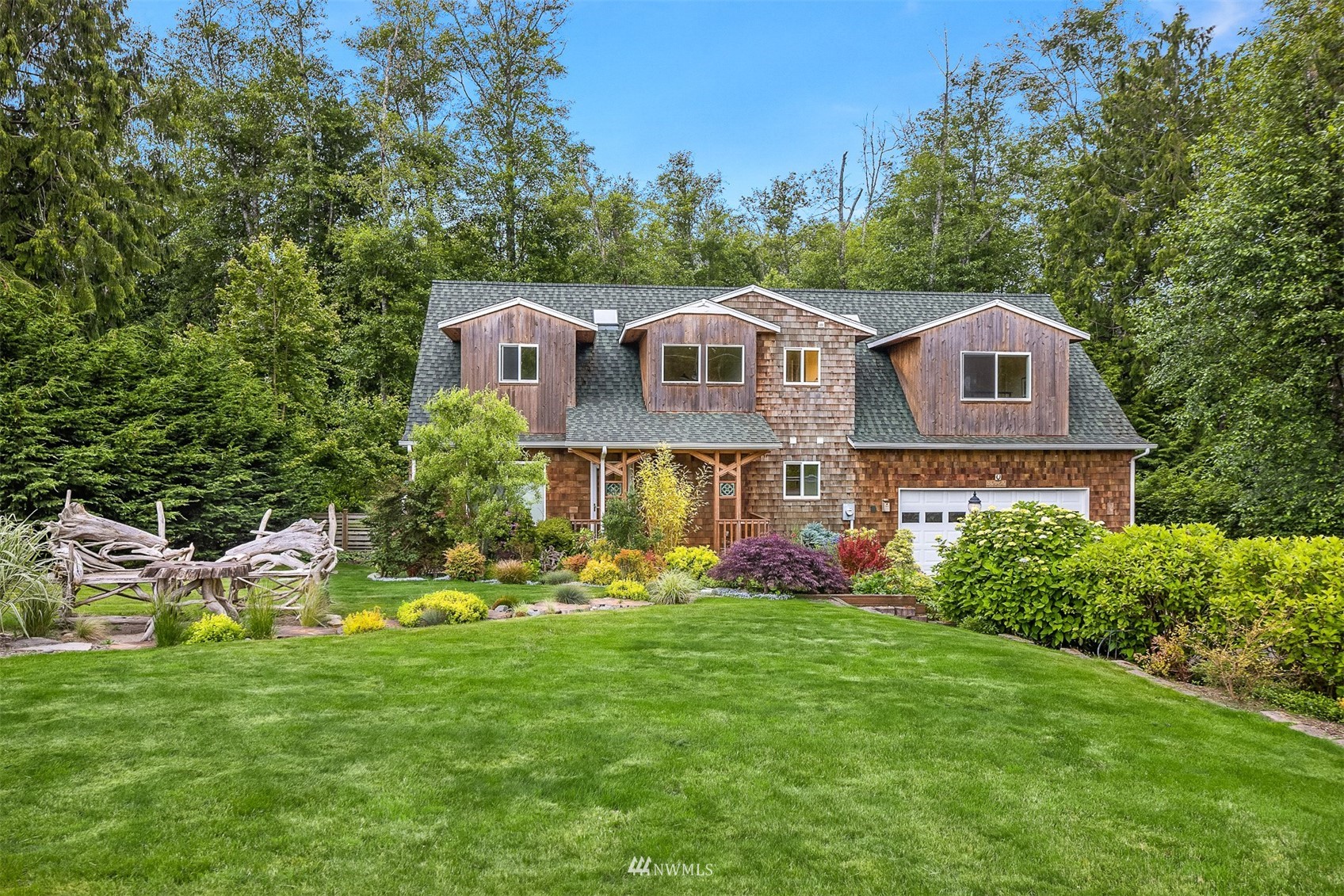 Cedar-shingle home with three dormers, attached garage, landscaped lawn, and wooded surroundings.