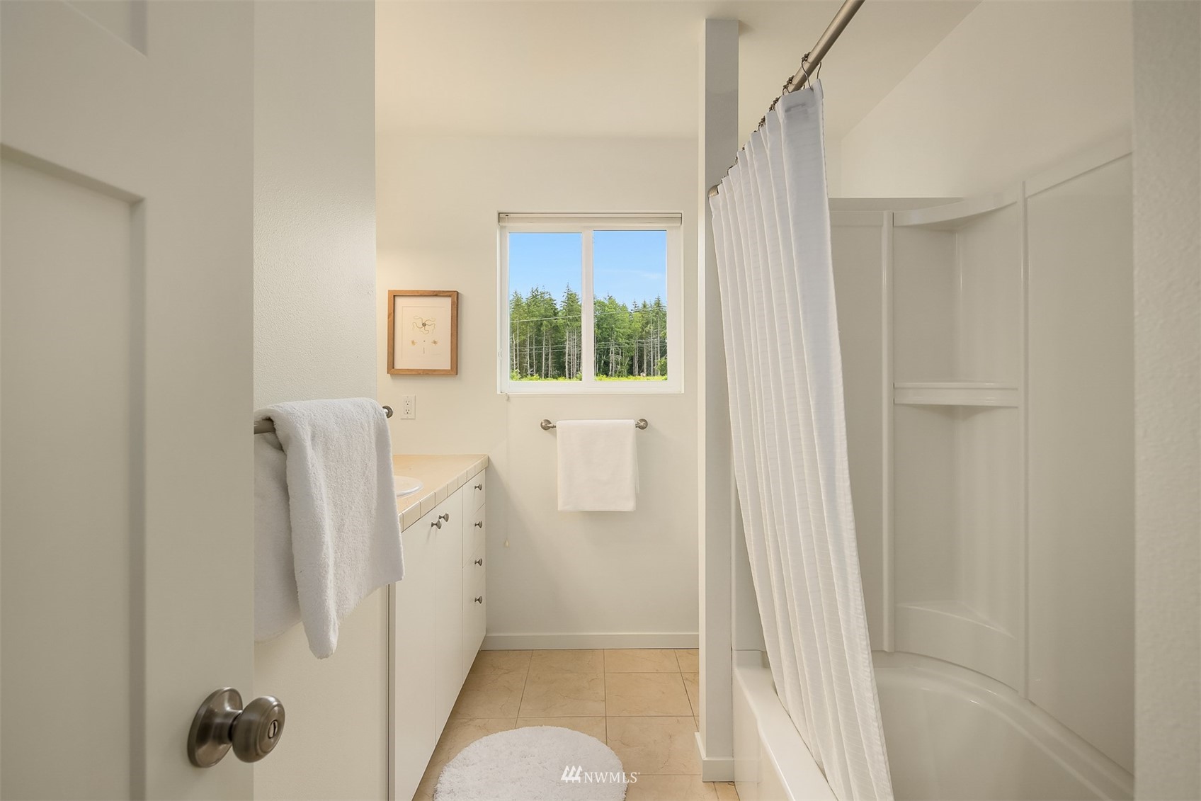 Bathroom with a white vanity, tiled floor, and curtained tub beside a window overlooking trees.