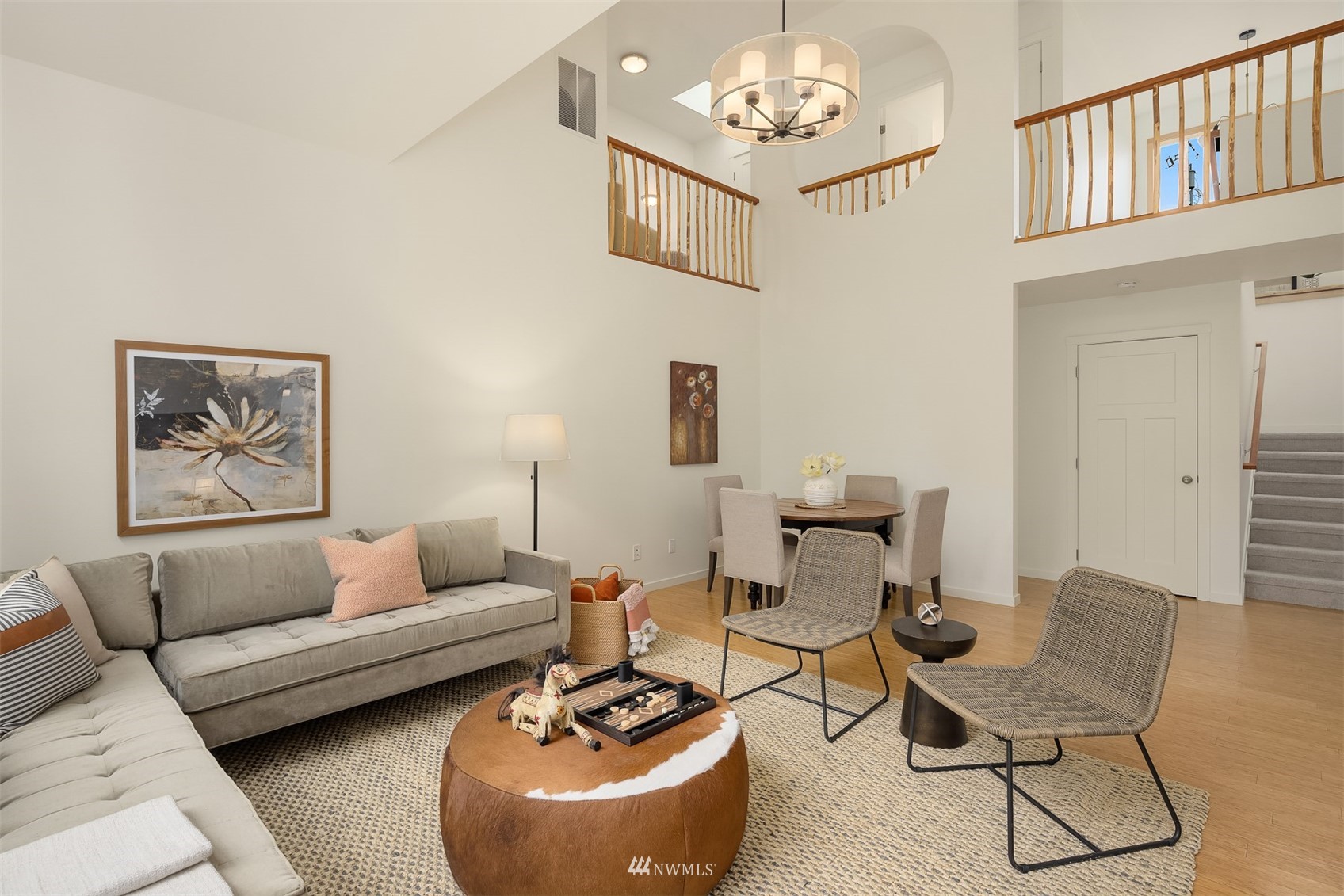Double-height living room with gray sectional, woven chairs, and an open dining area beneath an upstairs landing.