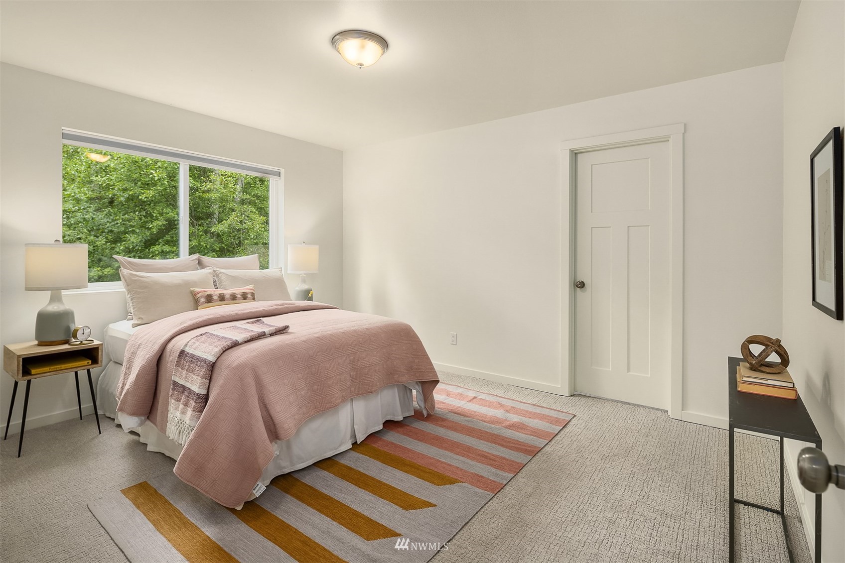 Staged bedroom with blush-pink bedding, a striped rug, and a large window overlooking greenery.