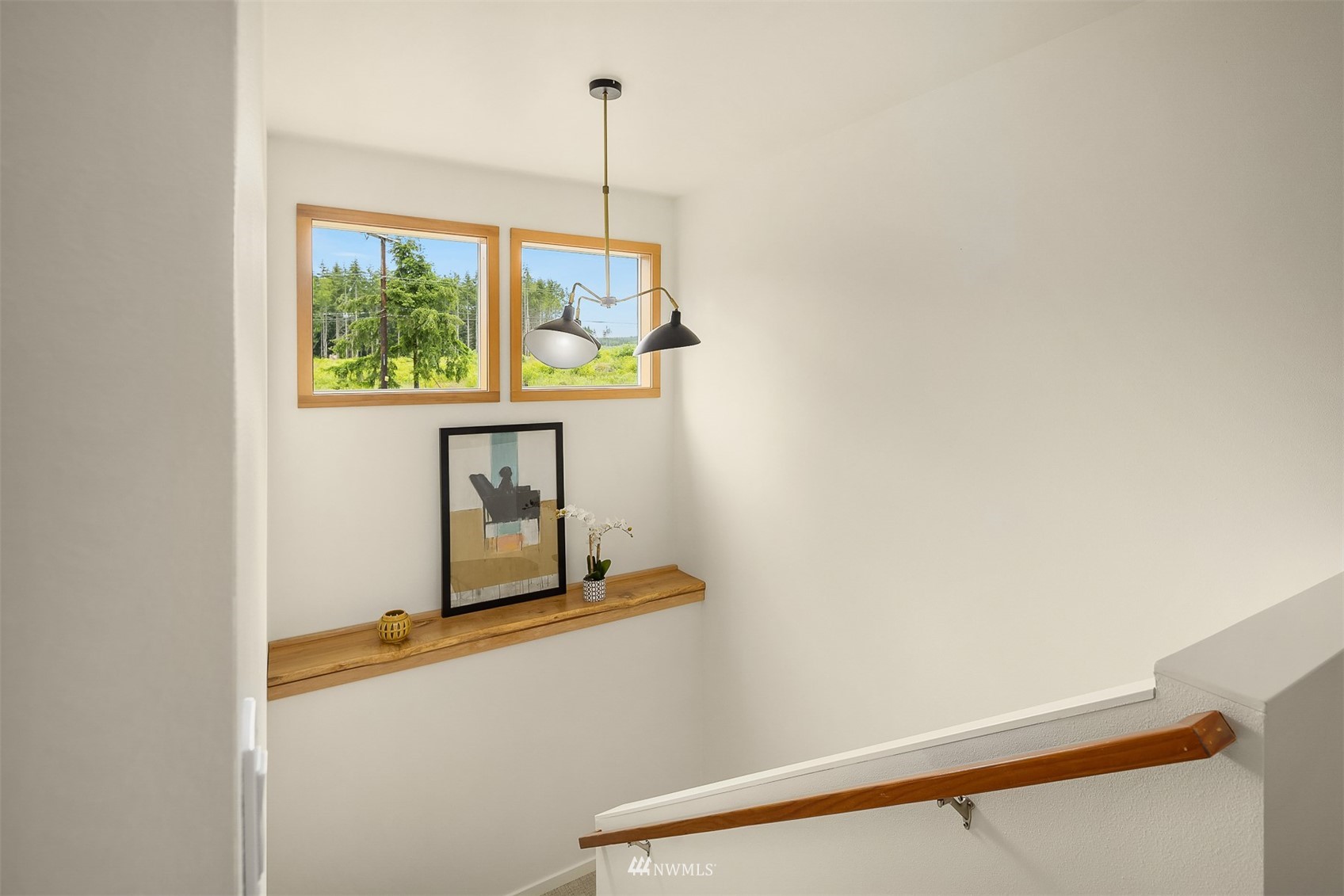 Stairwell with a wooden handrail, two windows overlooking greenery, and a shelf with artwork and a plant.