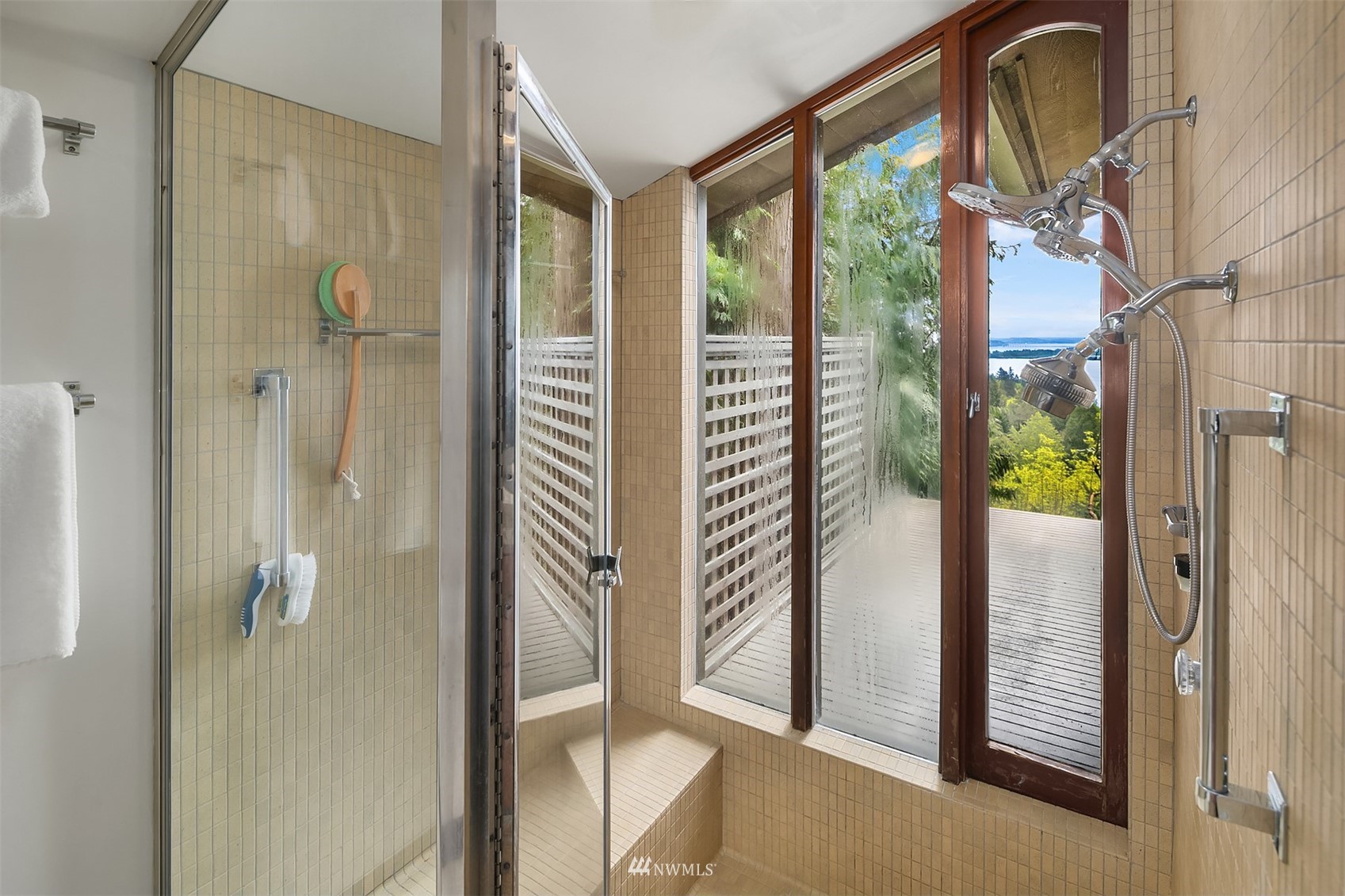 Tiled bathroom shower with multiple showerheads and windows overlooking a deck and distant water.