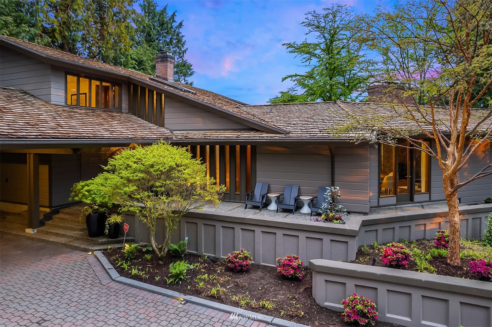 Modern gray home with a lit patio, layered shingle roofs, and flowering landscaping at dusk.