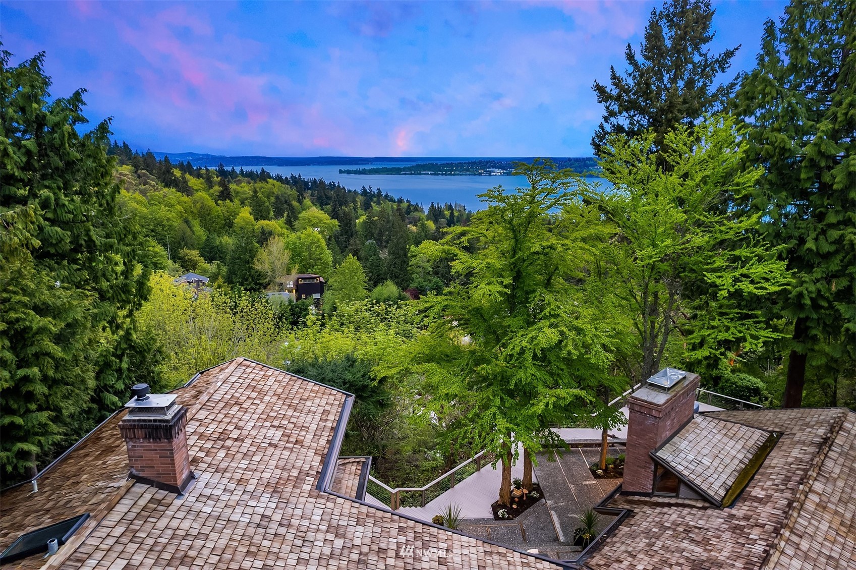 Home rooftops and landscaped deck frame a lake view beyond dense green trees.