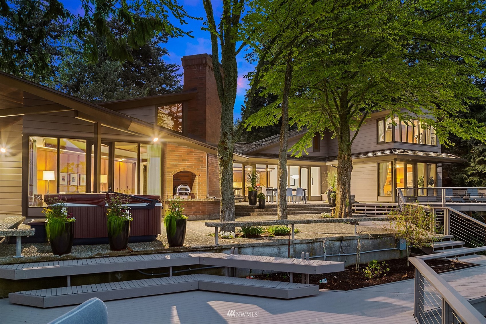 Modern house at dusk with a brick chimney, hot tub, and broad deck beneath mature trees.
