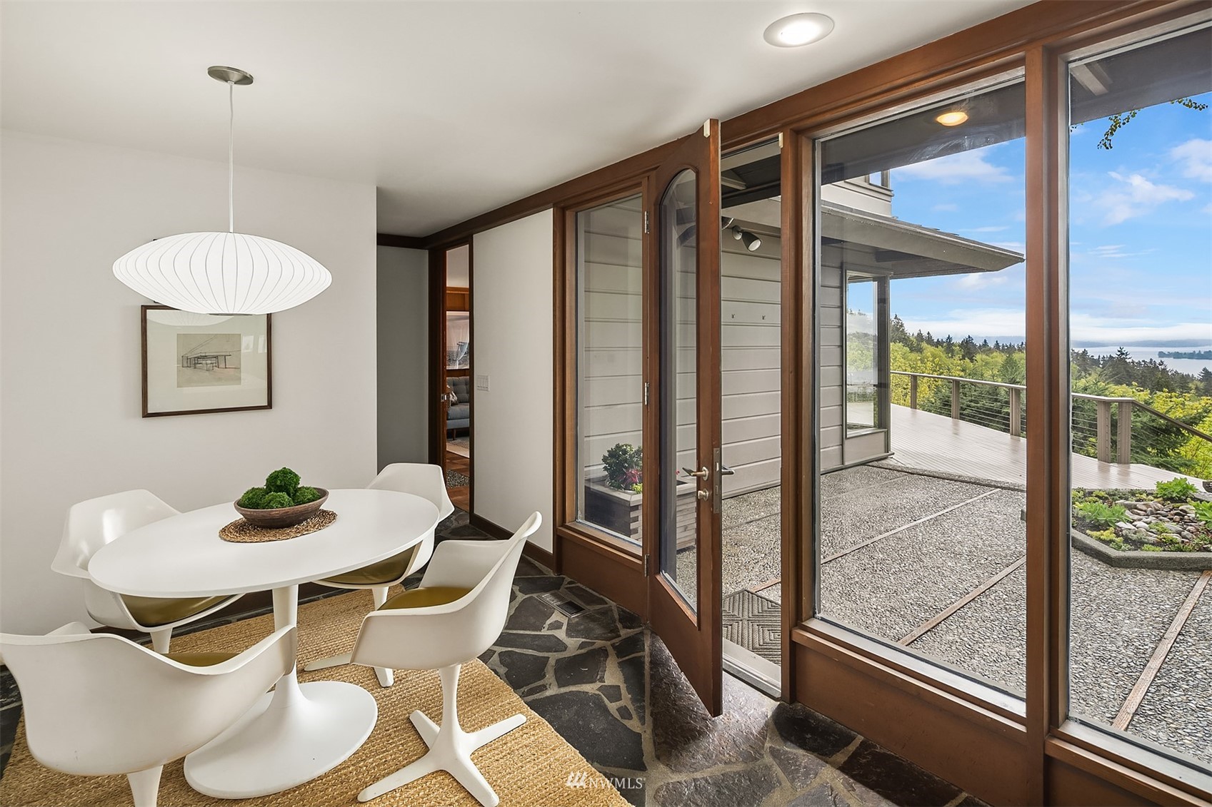 Dining room with a white table and chairs beside glass doors opening to a deck with distant water views.