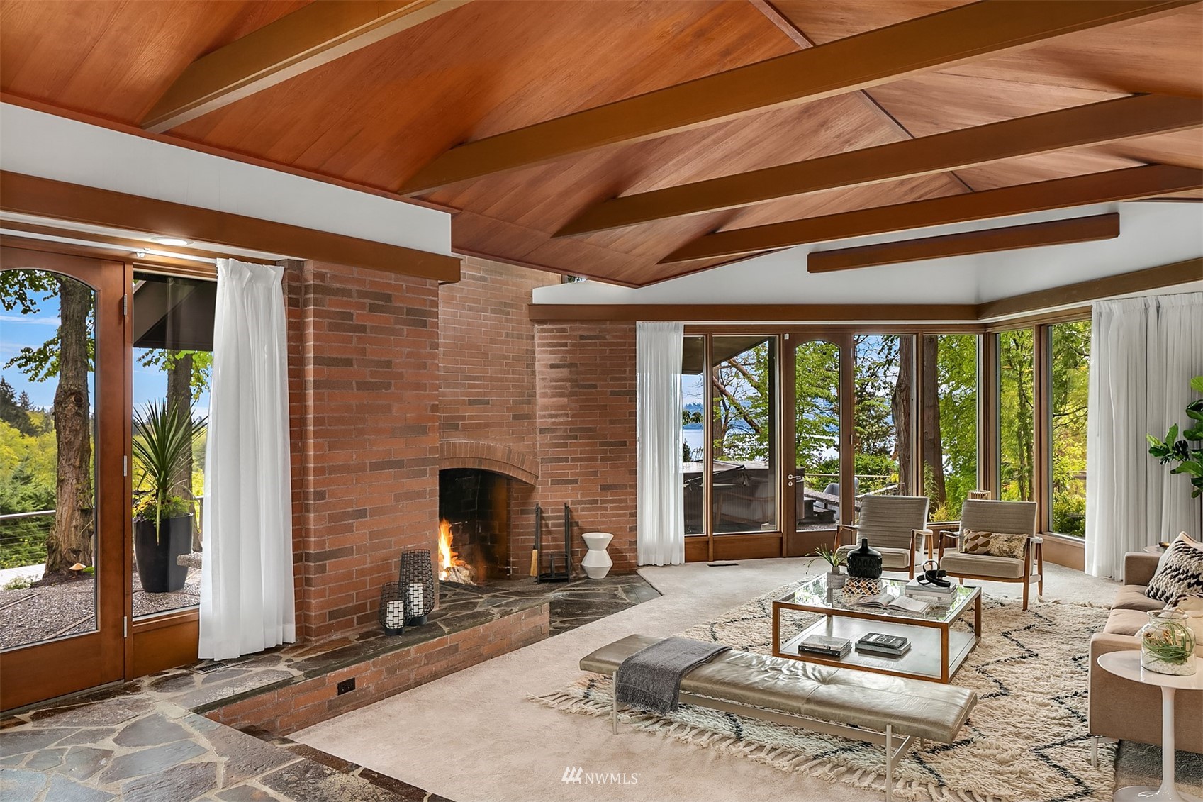 Living room with a vaulted wood ceiling, brick fireplace, and large windows overlooking water.