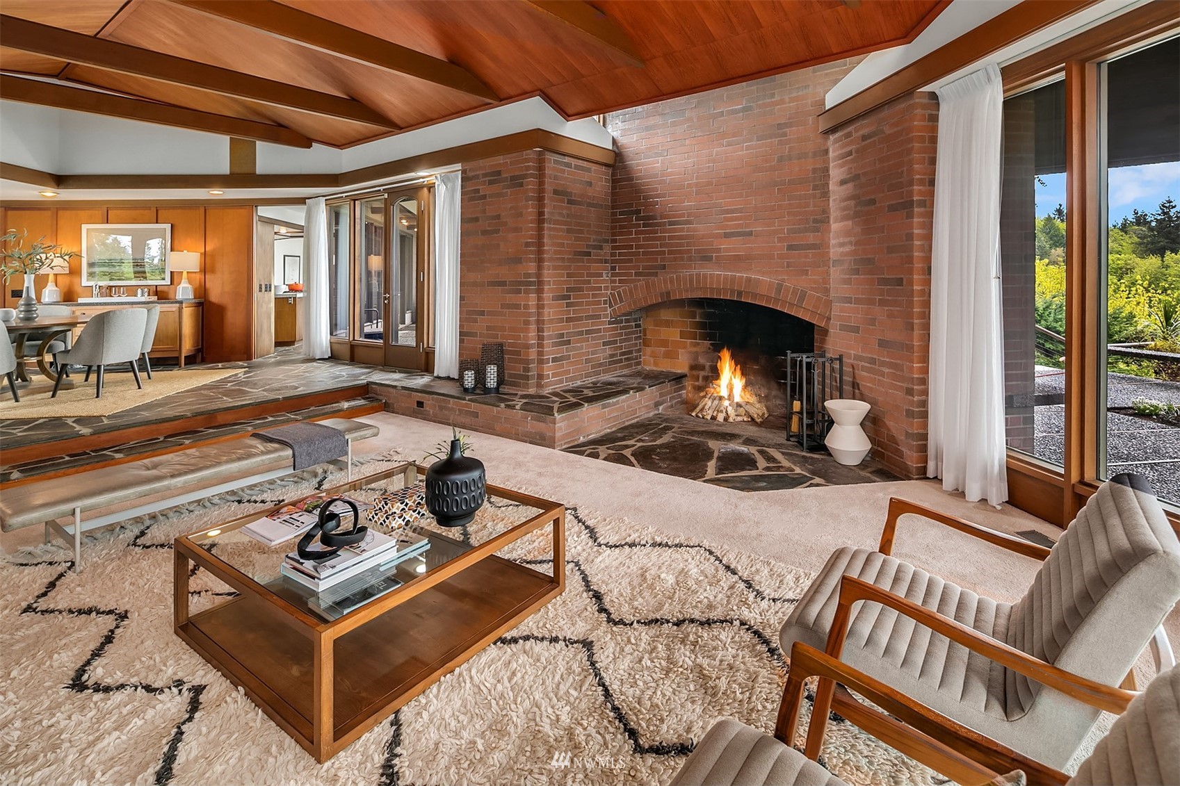 Living room with a brick fireplace, vaulted wood ceiling, and large windows.