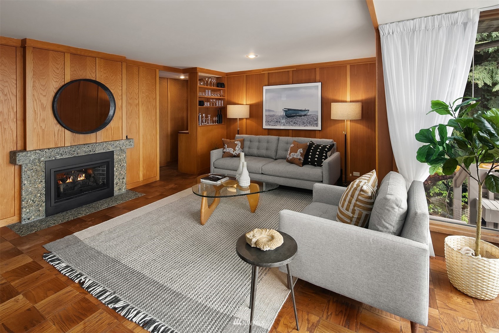 Living room with wood-paneled walls, a stone fireplace, gray sofas, and a large window overlooking greenery.
