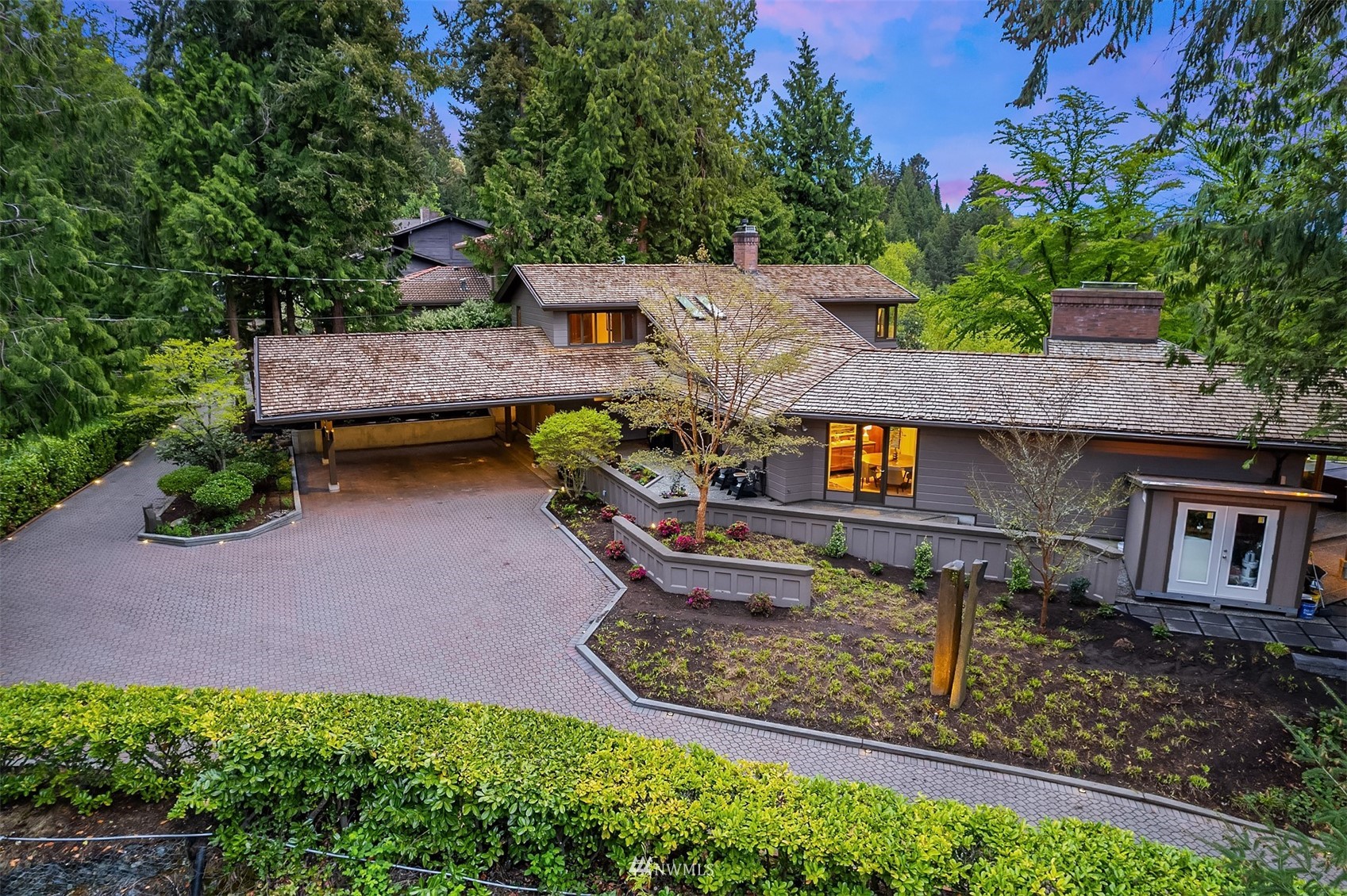 Large gray house with a shingled roof, paved driveway, and landscaped gardens among evergreen trees.