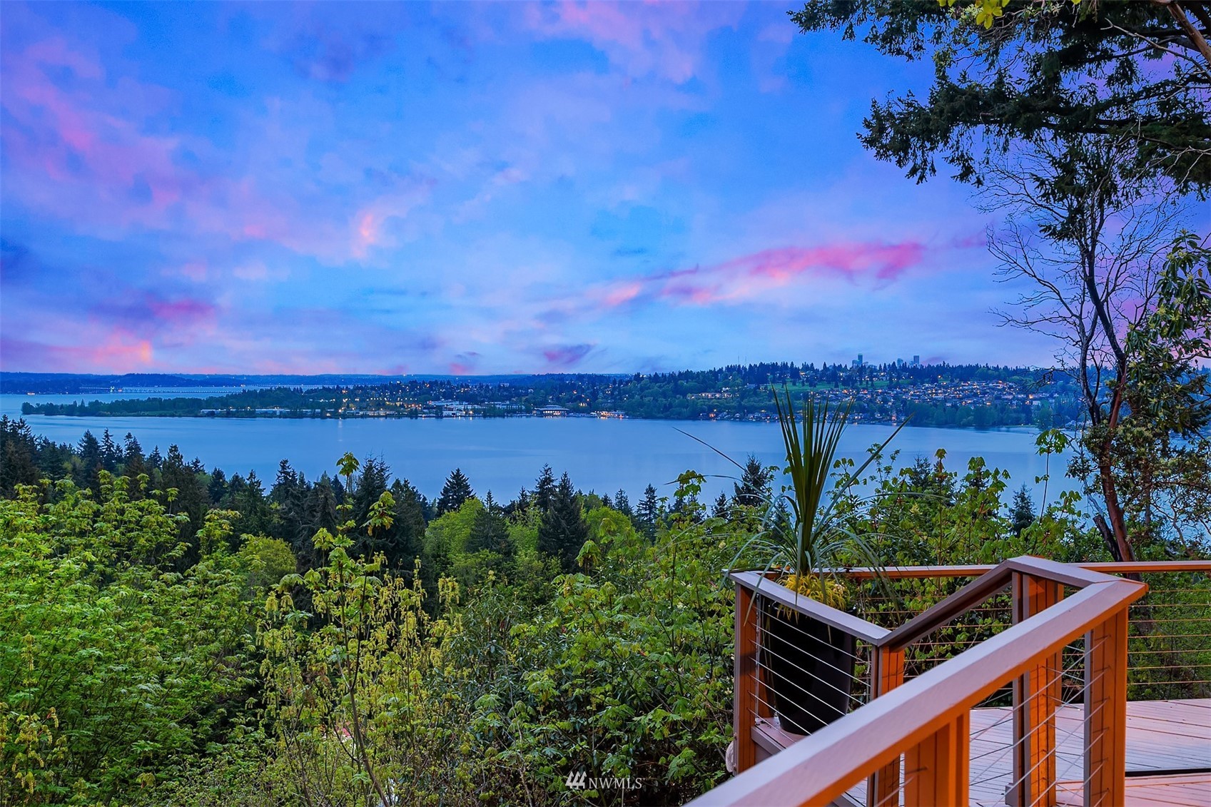 Water and distant city at dusk, framed by trees and a deck railing.