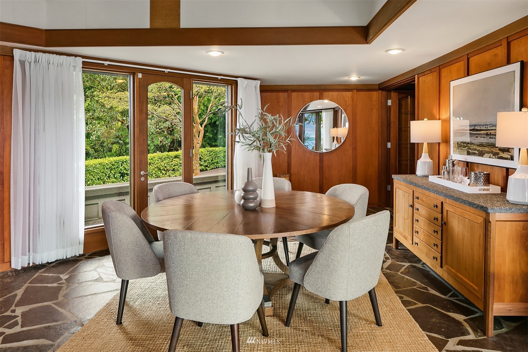 Dining room with a round wooden table, wood-paneled walls, and glass doors overlooking a leafy garden.