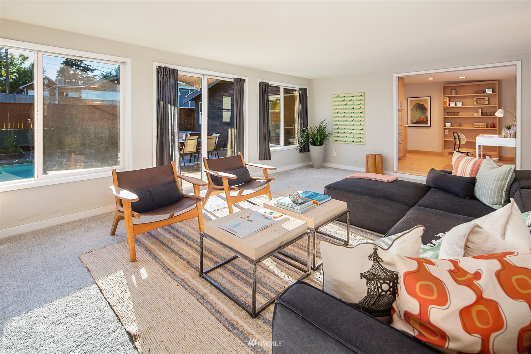 Living room with a dark sectional, wood lounge chairs, and large windows overlooking a backyard pool.