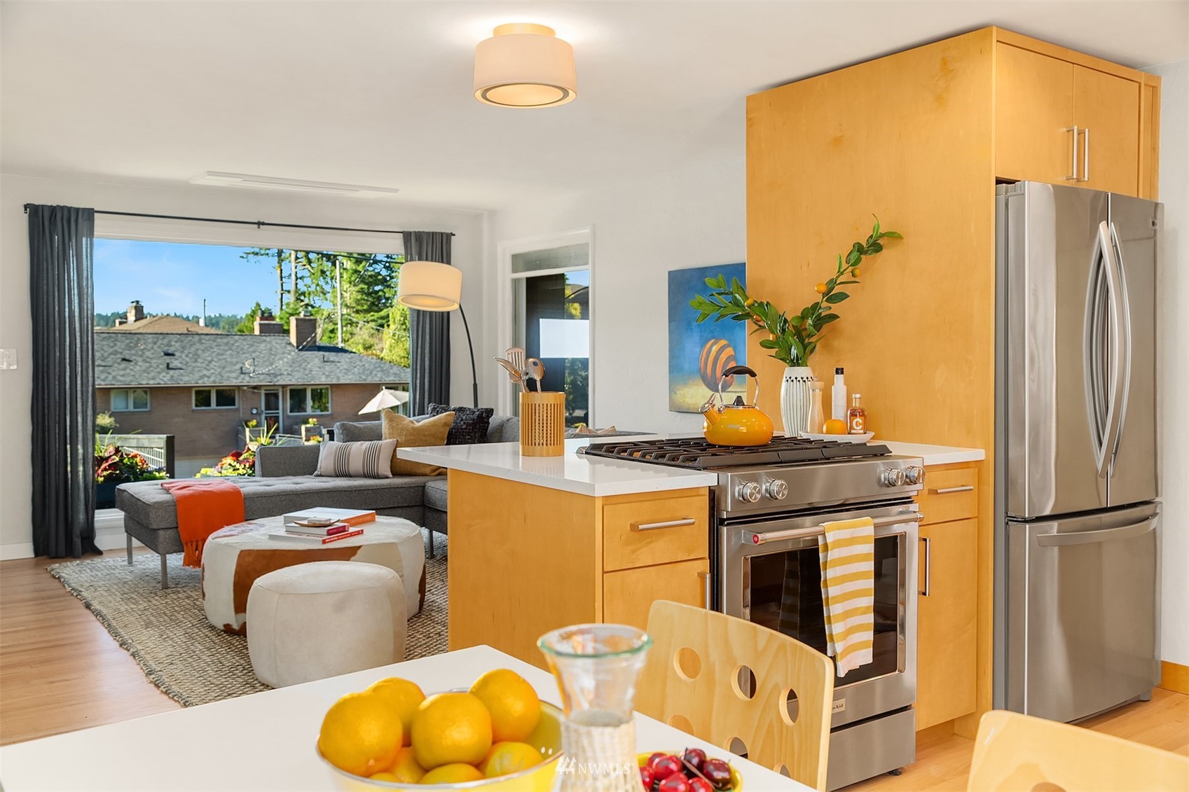 Open-plan kitchen with wood cabinets and stainless appliances beside a living room with a gray sectional.
