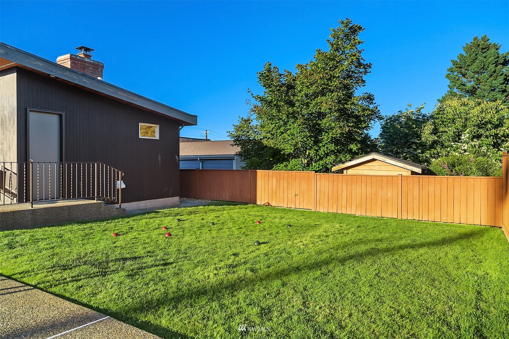 Fenced backyard with a broad lawn, colored game balls, mature trees, and a small shed.