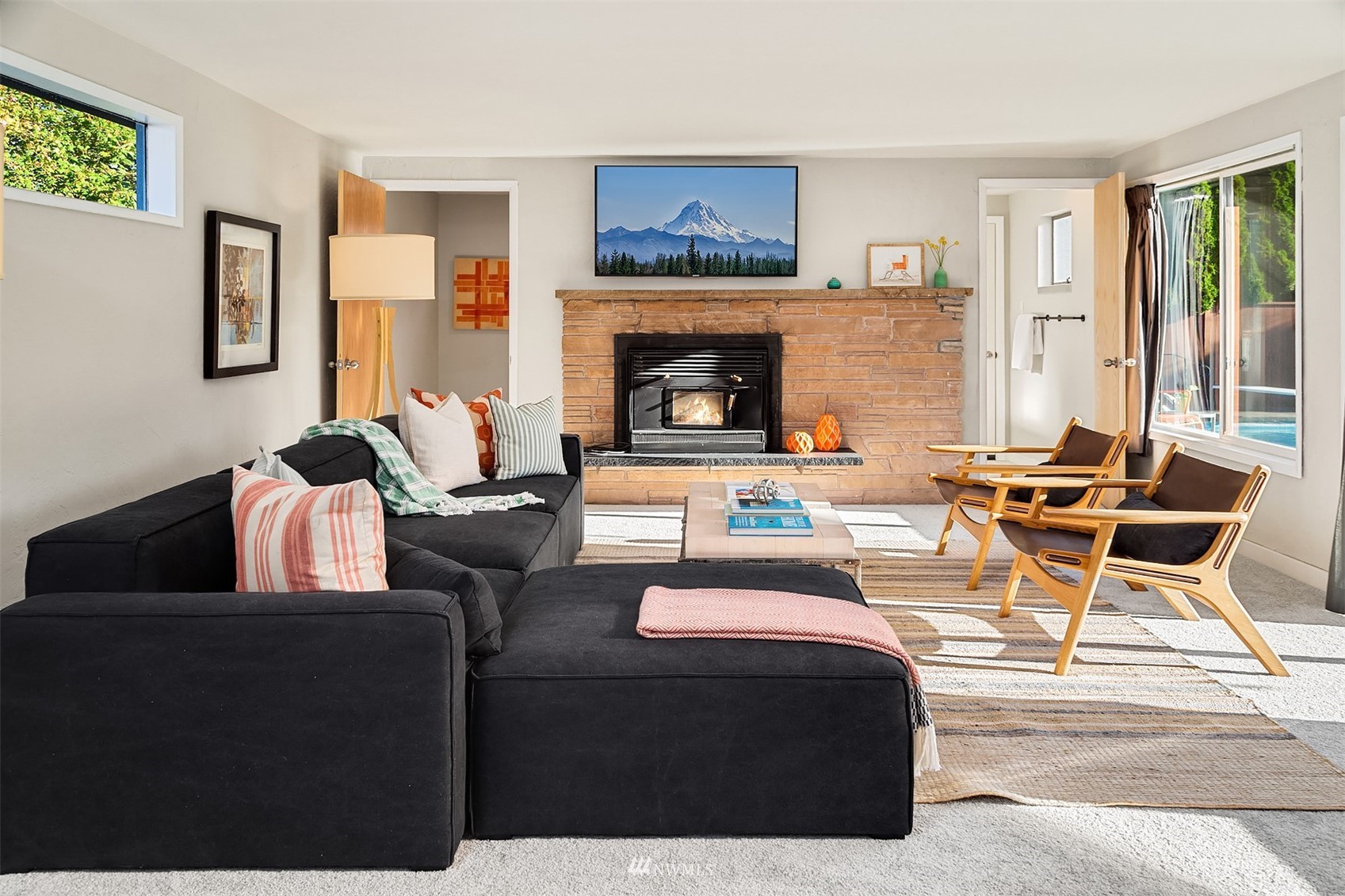 Living room with a stone fireplace, dark sectional, wooden lounge chairs, and pool visible through the windows.