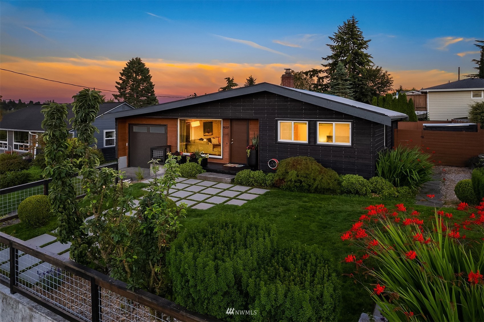 Single-story black house with warm lit windows, a landscaped garden, and a grid-pattern walkway at dusk.