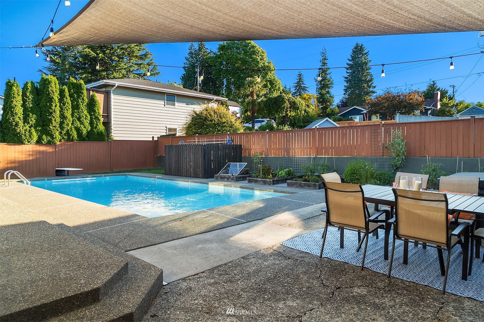 Backyard swimming pool beside a furnished patio, garden beds, and a tan shade sail.