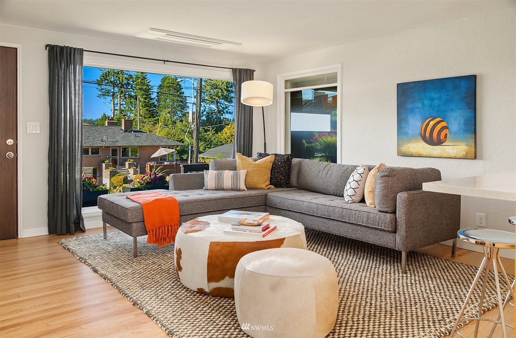 Staged living room with a gray sectional, round ottomans, hardwood floors, and a large window.
