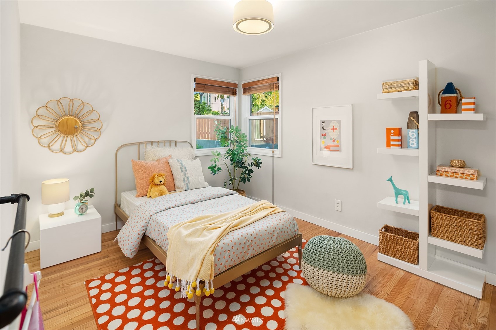 Staged bedroom with a brass bed, dotted orange rug, white shelving, and two corner windows.