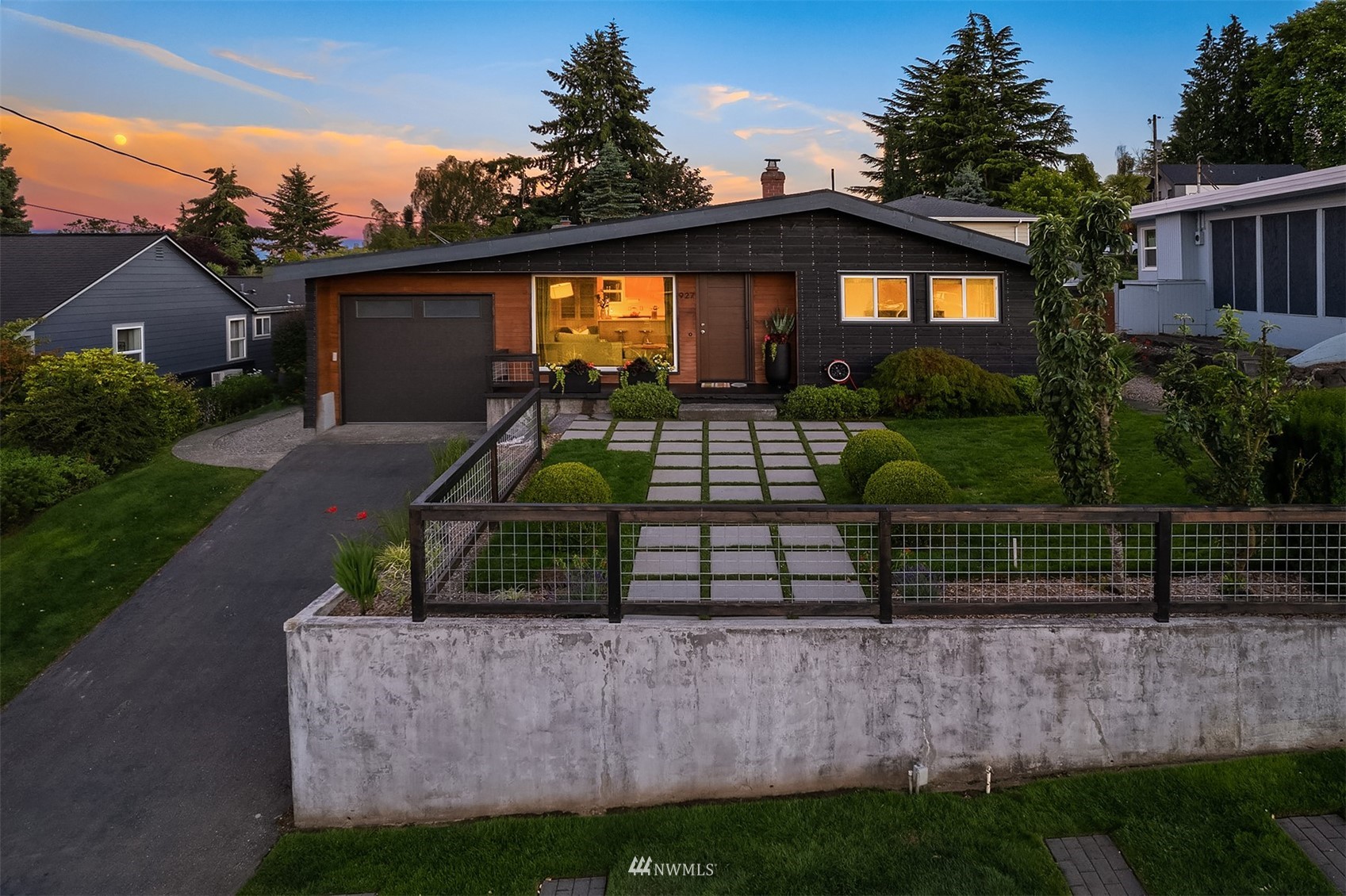 Single-story home with dark siding, wood accents, landscaped yard, and paved path at sunset.