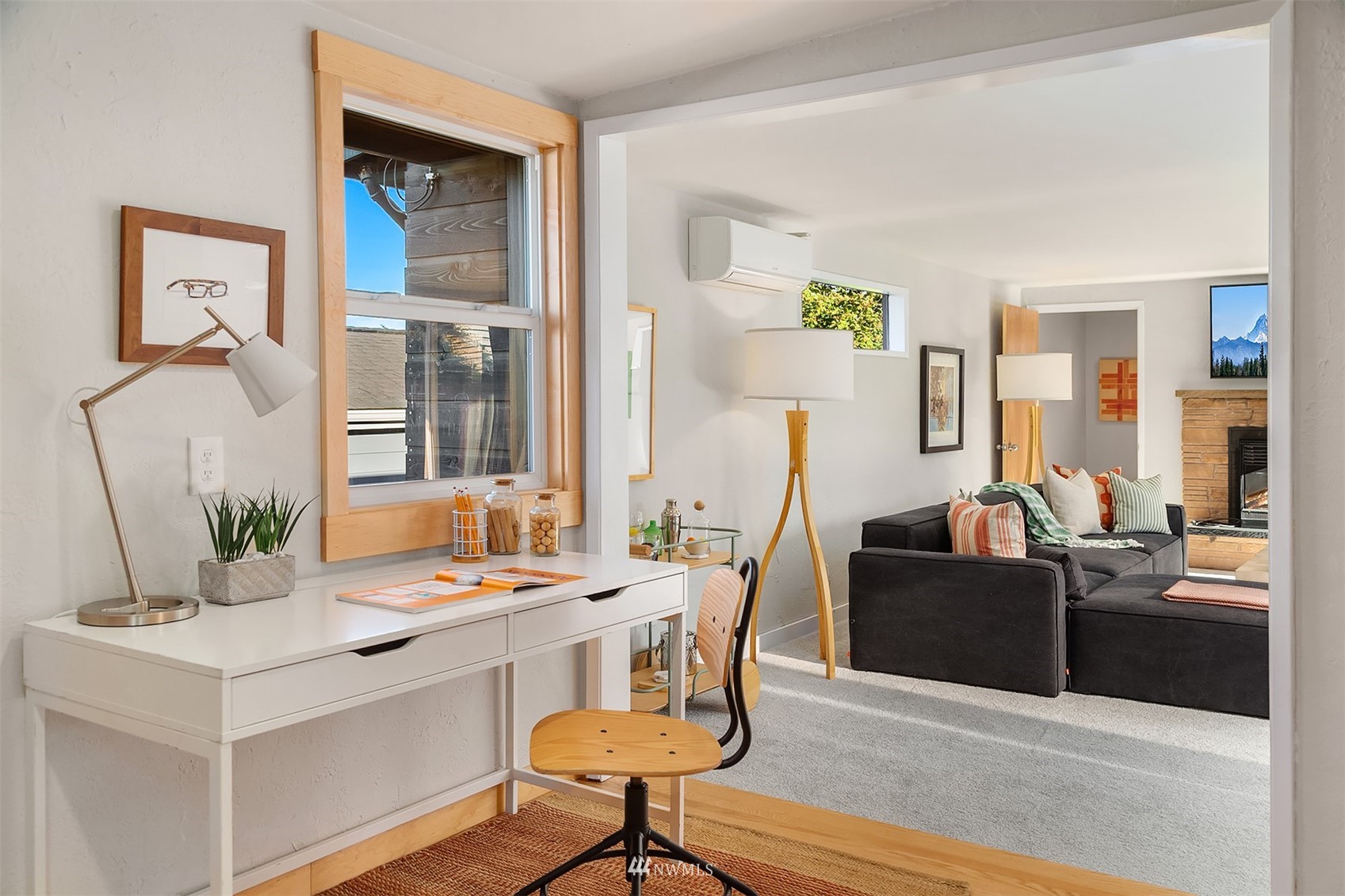 Home office nook with a white desk opens into a living room with a dark sectional and stone fireplace.