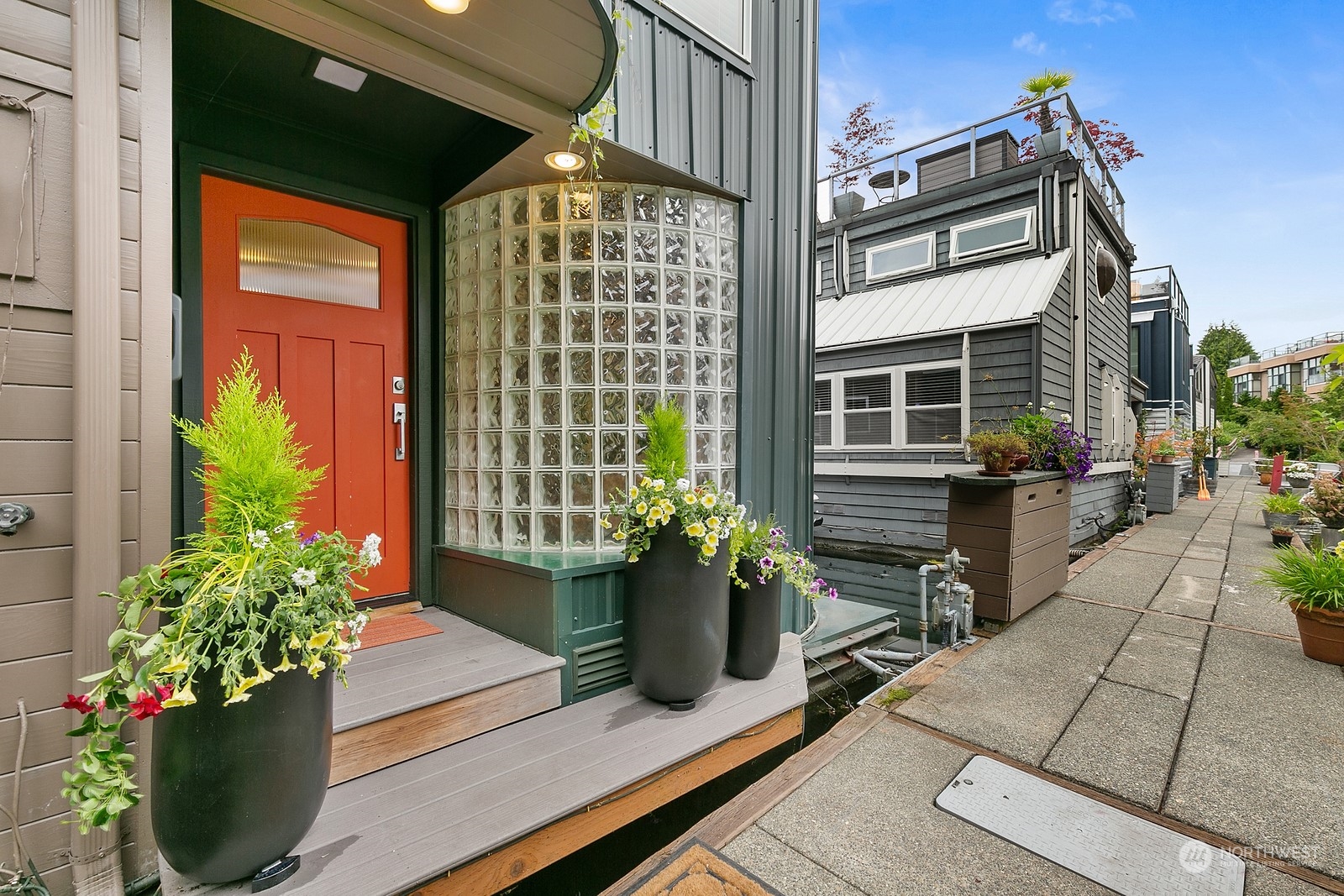 Modern home entrance with a red door, curved glass-block wall, and flower-filled planters beside a paved walkway.