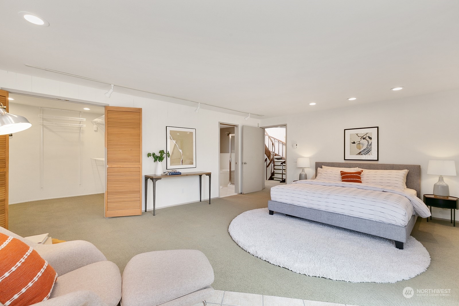 Staged bedroom with a gray bed, open closet with wooden doors, and a staircase visible through the doorway.